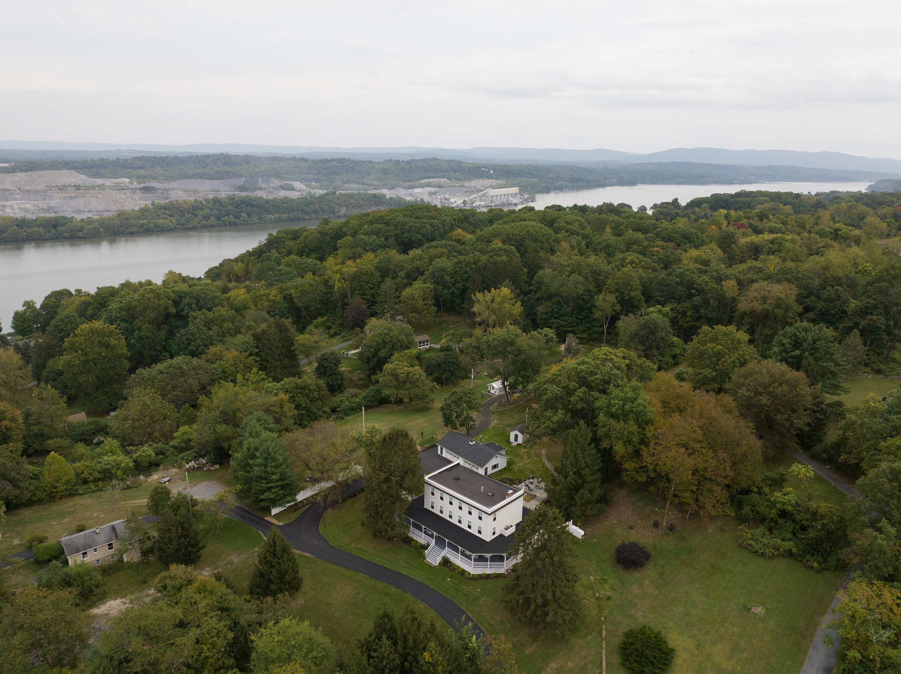 A wide aerial shot showcasing the property nestled among dense green trees, with a river and industrial structures visible in the distance. The large white house with its dark roof and surrounding greenery dominates the foreground.