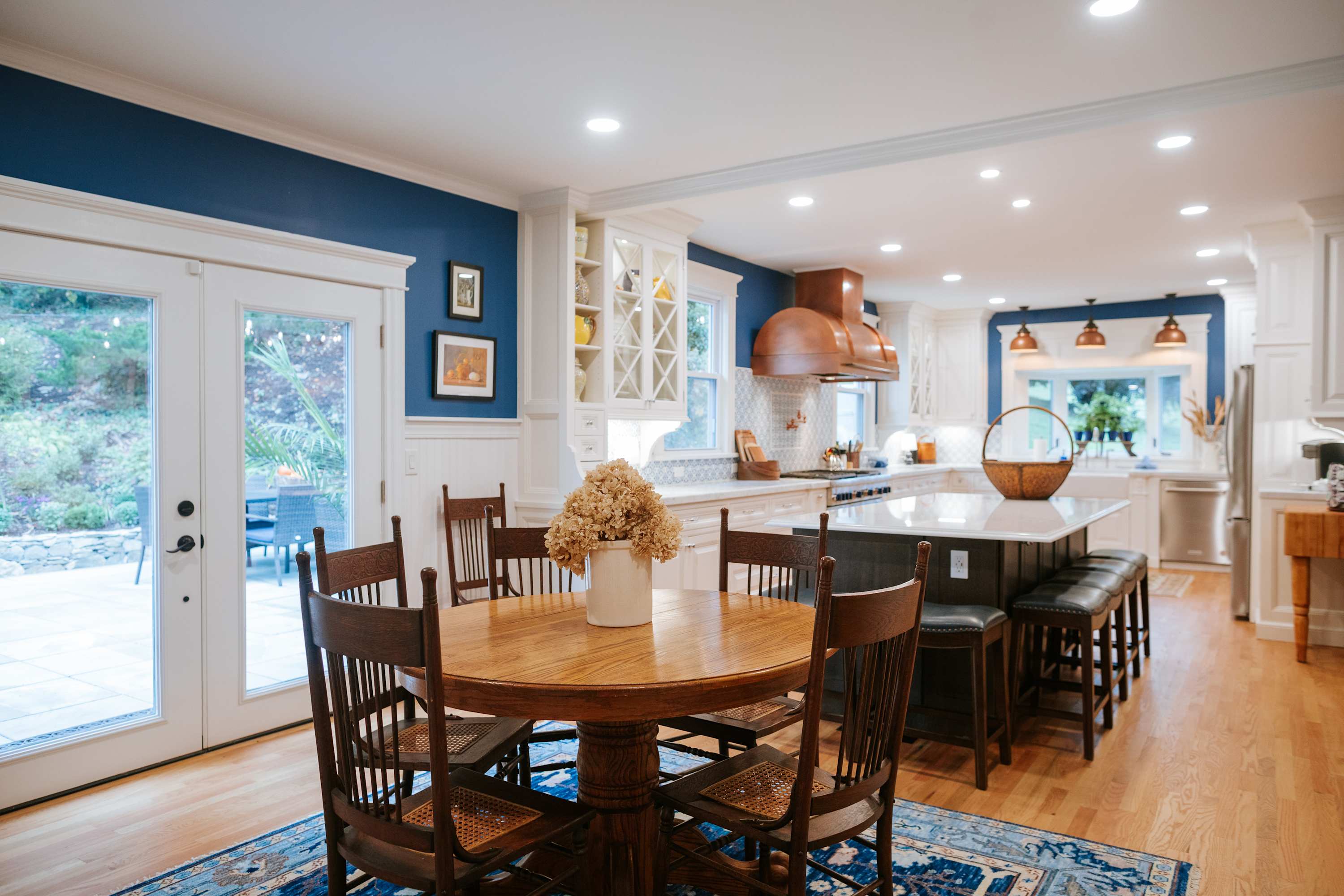Spacious kitchen with a dining nook, white cabinetry, a large island with seating, stainless steel appliances, and a copper range hood, featuring blue accent walls and hardwood floors.
