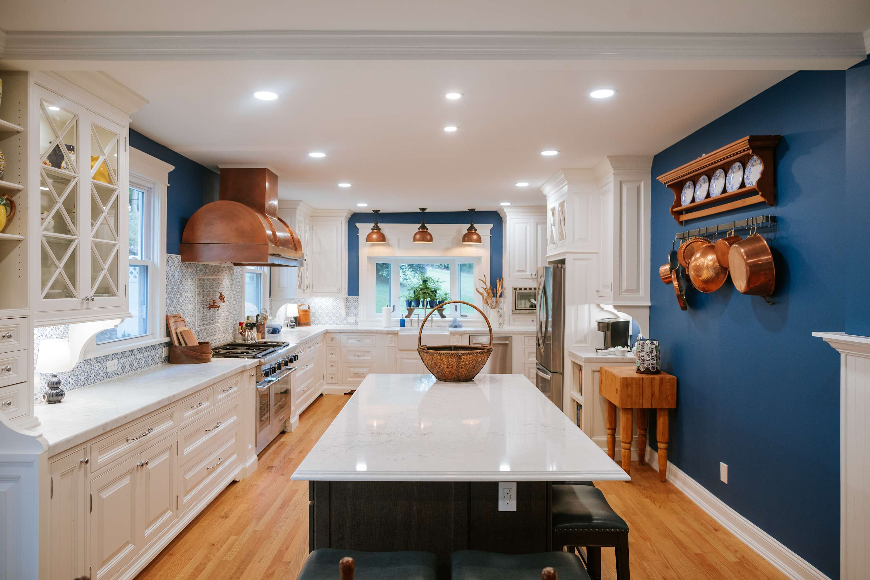 Bright and spacious kitchen featuring white cabinetry, marble countertops, a large island with seating, and a striking blue accent wall. Copper accents are visible in the range hood and hanging pots.