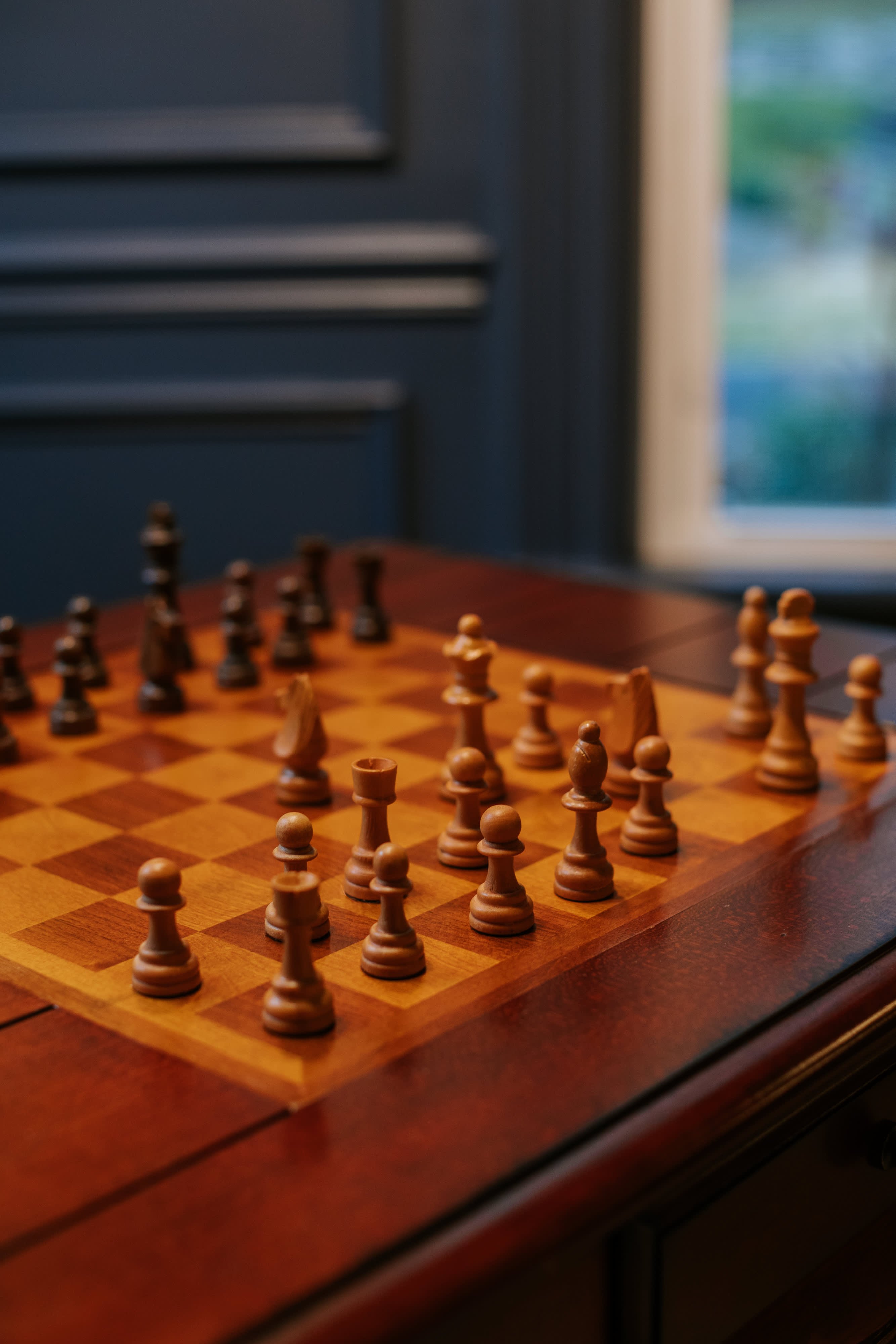 Close-up of a wooden chess set arranged on a polished wooden game table, with a blurred background showing a window and dark paneled walls.