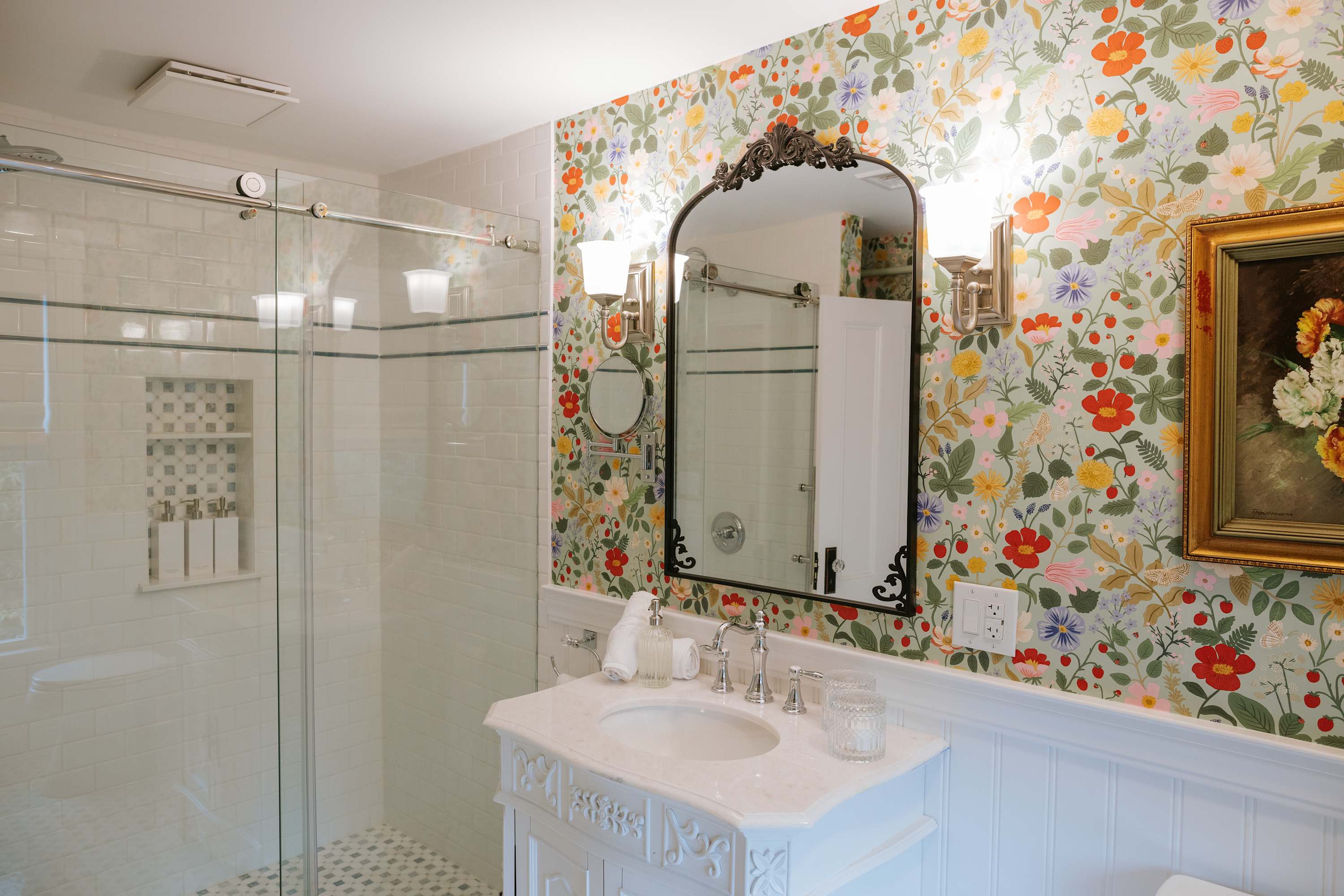 A bright bathroom featuring floral wallpaper, white wainscoting, a white vanity with a marble top, and a glass-enclosed shower with white subway tiles and a mosaic floor. An ornate mirror and wall sconces are visible.