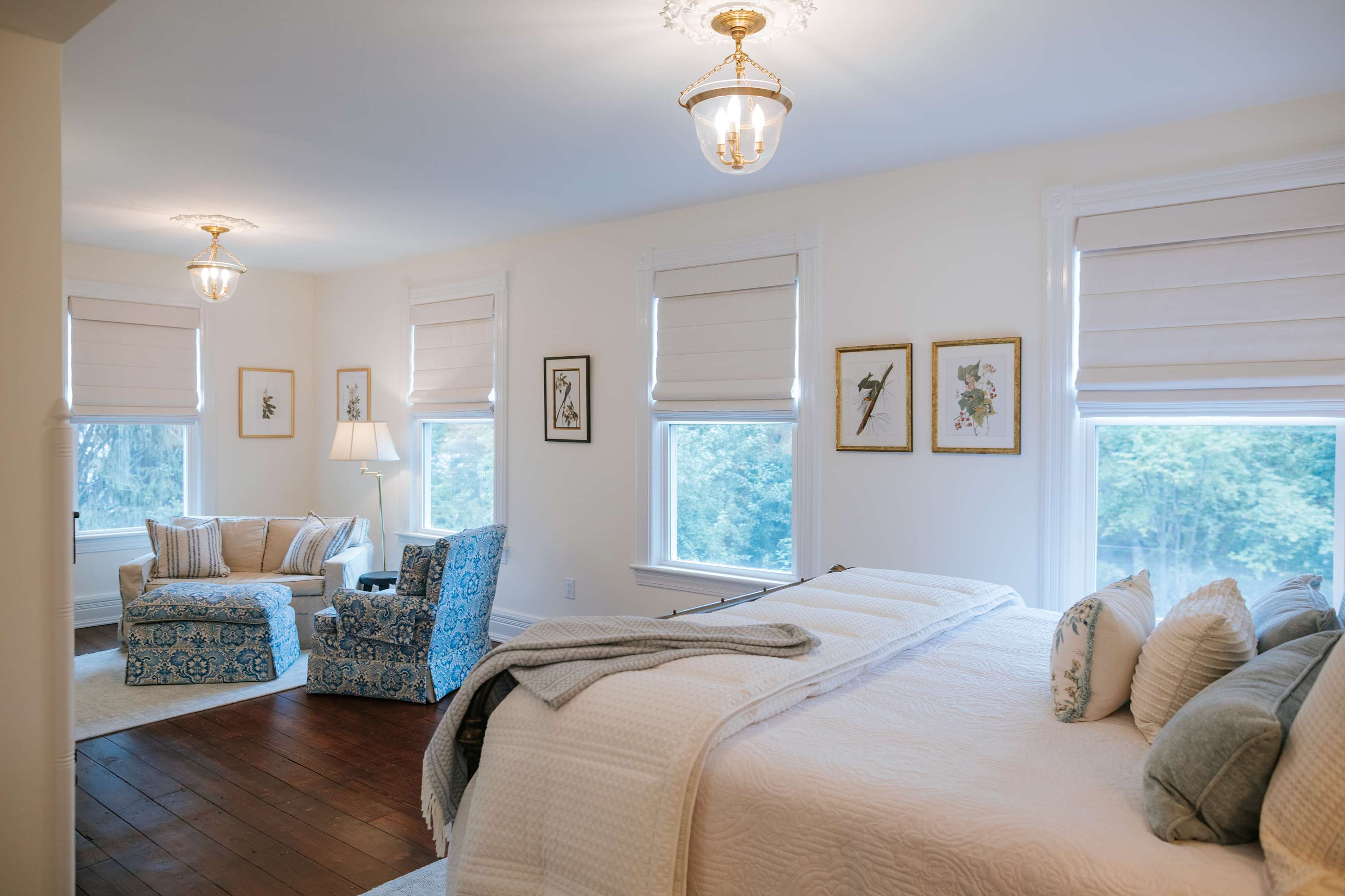 A spacious bedroom featuring a large bed with white bedding and blue accents, dark hardwood floors, and a sitting area with a cream sofa and two blue patterned armchairs. Multiple windows with Roman shades and decorative ceiling lights are present.