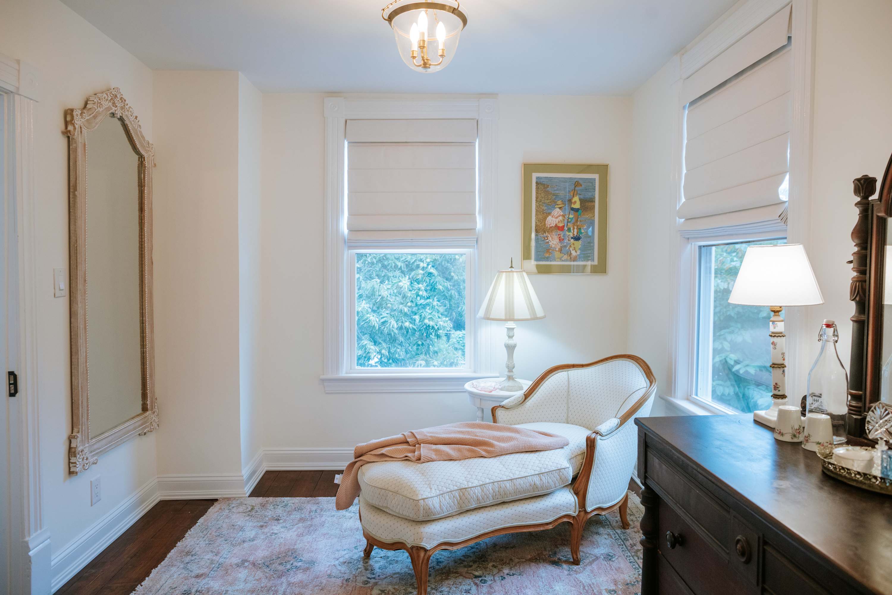 A bright and elegant bedroom featuring a white chaise lounge with a pink throw, a dark wood dresser with an antique mirror, and white walls. Two windows with Roman shades and a decorative ceiling light are visible.