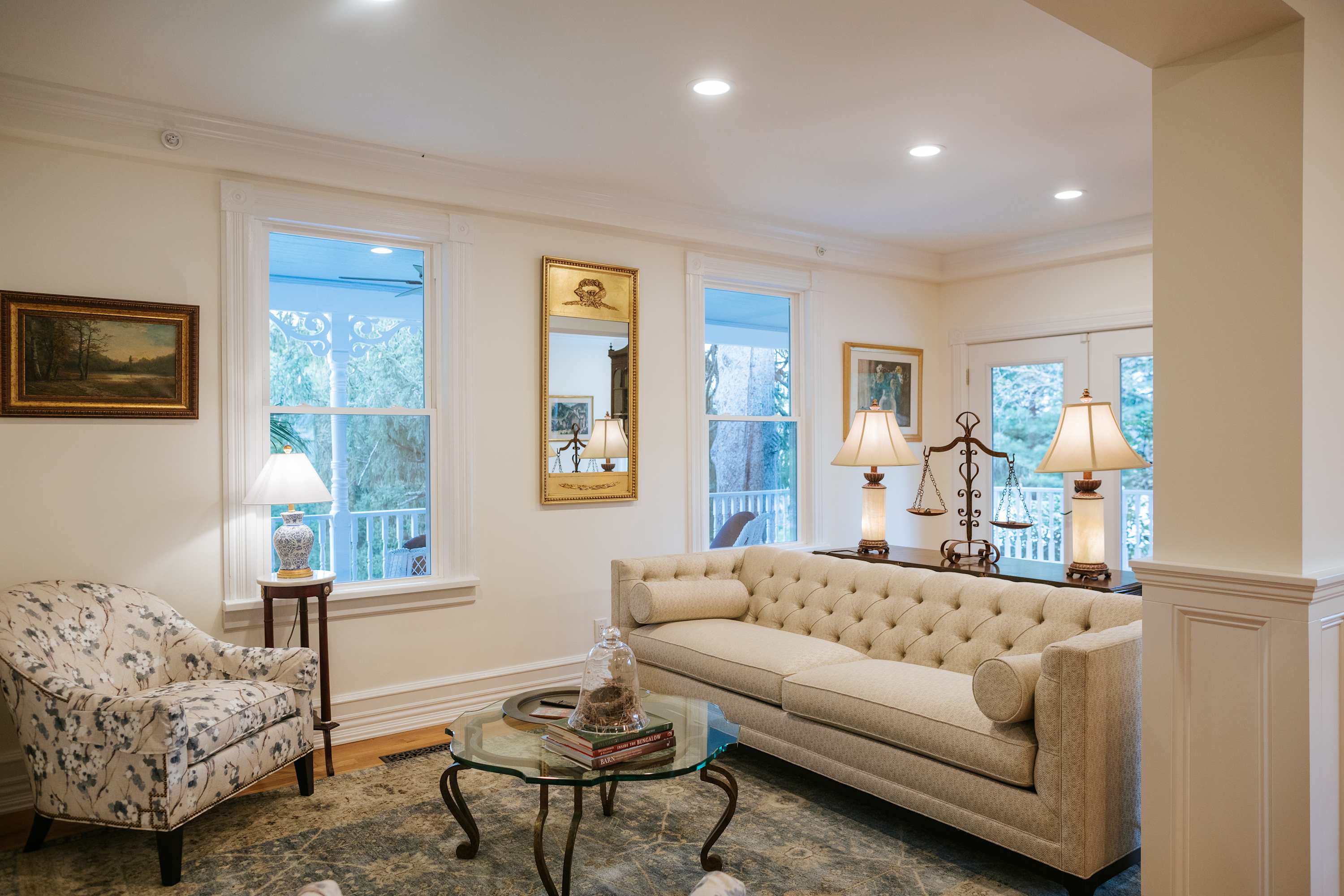 Elegant living room with a long tufted beige sofa, a floral armchair, a glass coffee table, and multiple windows, featuring a gold-framed mirror and decorative lamps.