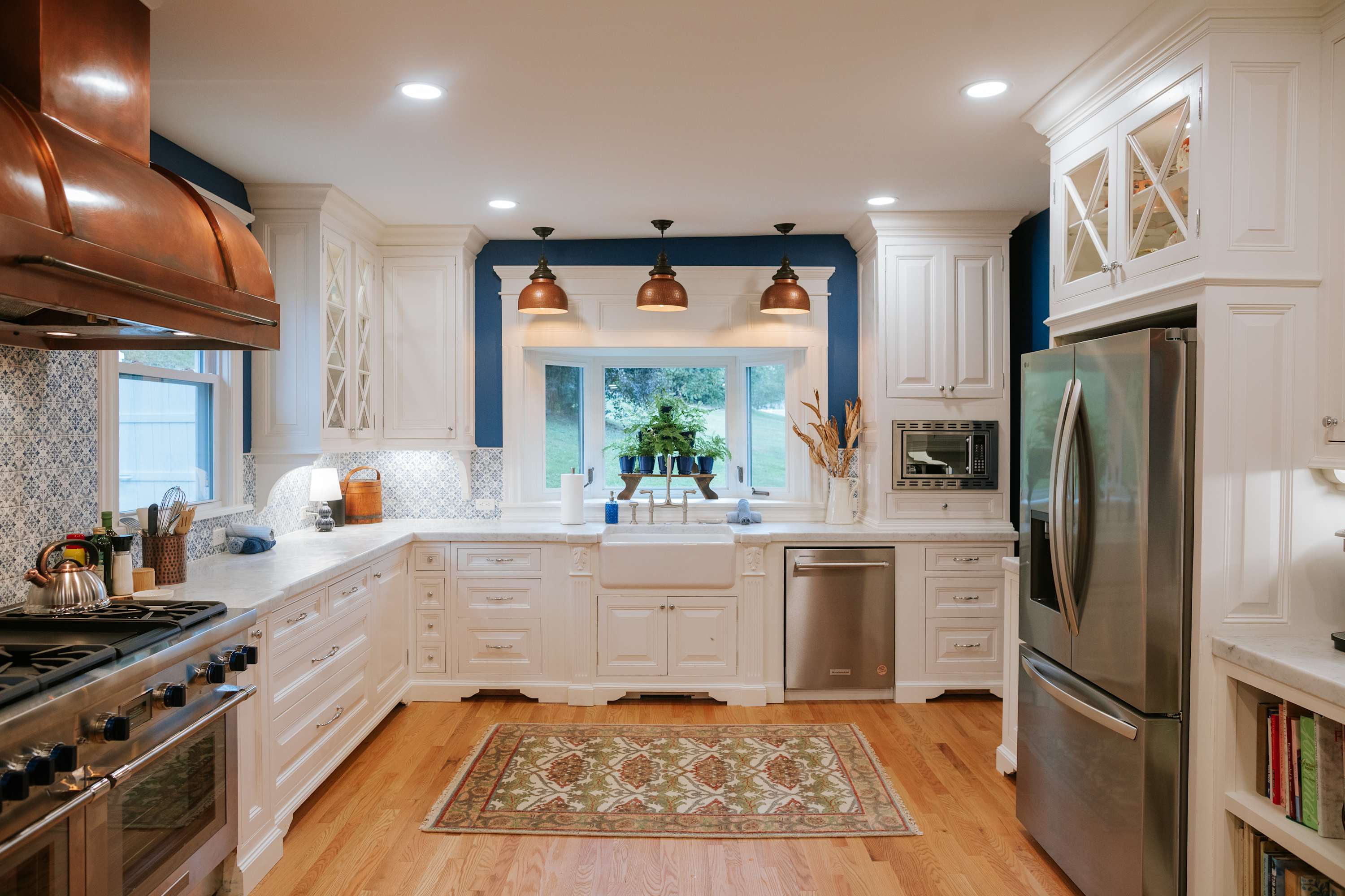 Bright kitchen with white shaker cabinets, marble countertops, a farmhouse sink, stainless steel appliances, and a decorative tile backsplash, featuring a large bay window and copper pendant lights.