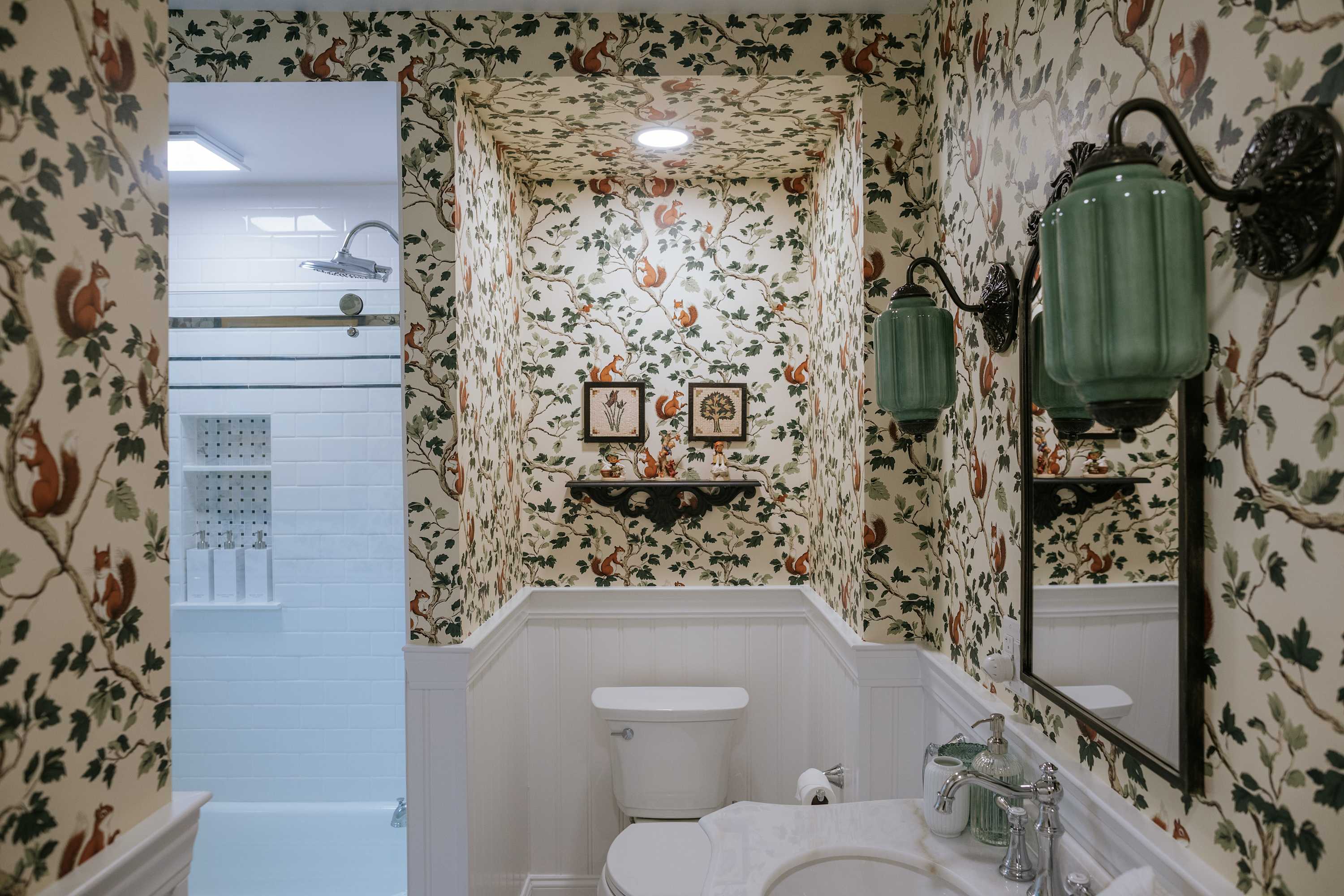 A whimsical bathroom featuring squirrel and foliage patterned wallpaper, white wainscoting, a white vanity with a marble top, and a toilet. A shower/tub combo with white subway tiles is visible in the background.