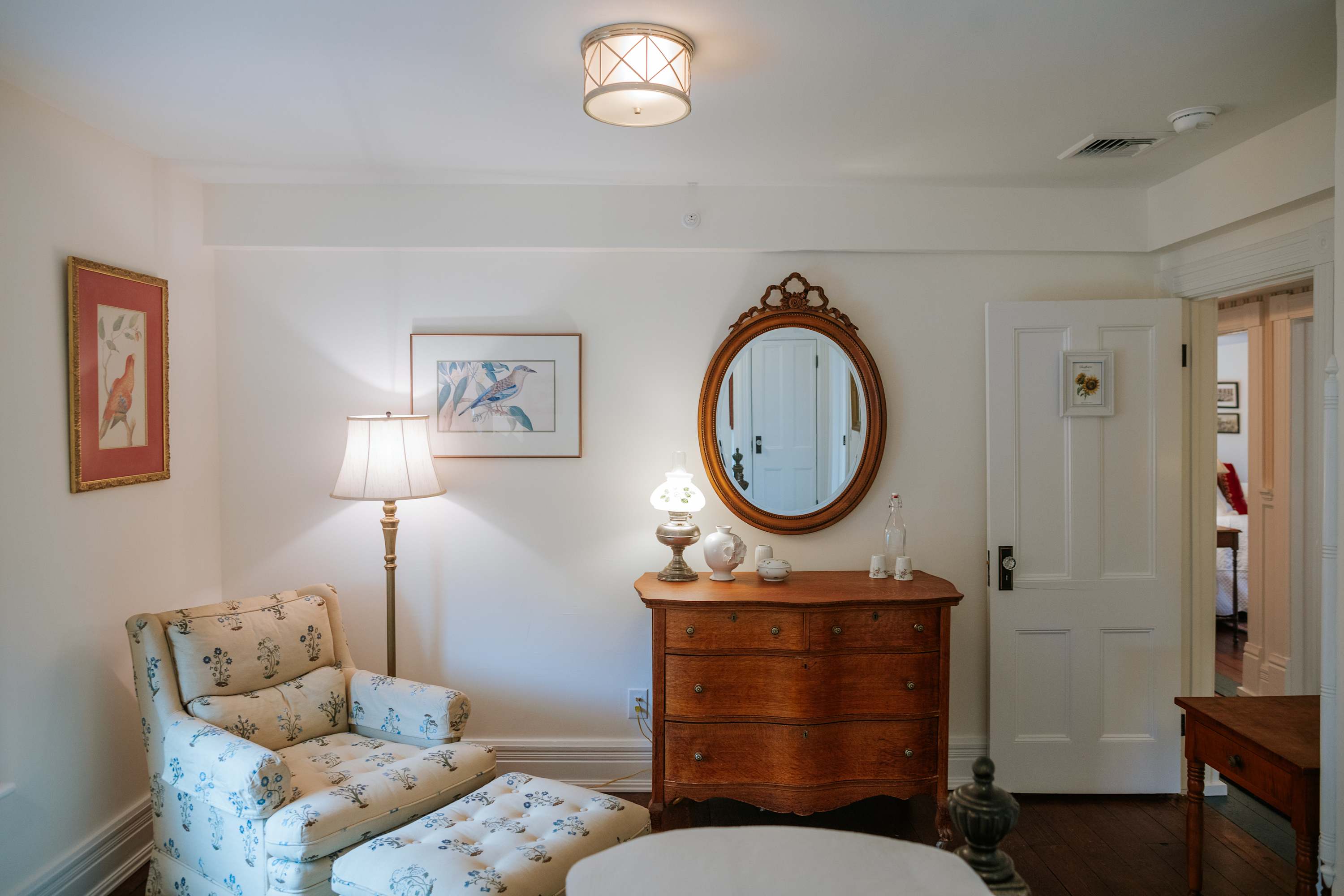 A cozy sitting area featuring a floral upholstered armchair with an ottoman, a wooden dresser with an oval mirror and a lamp, and framed artwork on the white walls, with a doorway leading to another room.