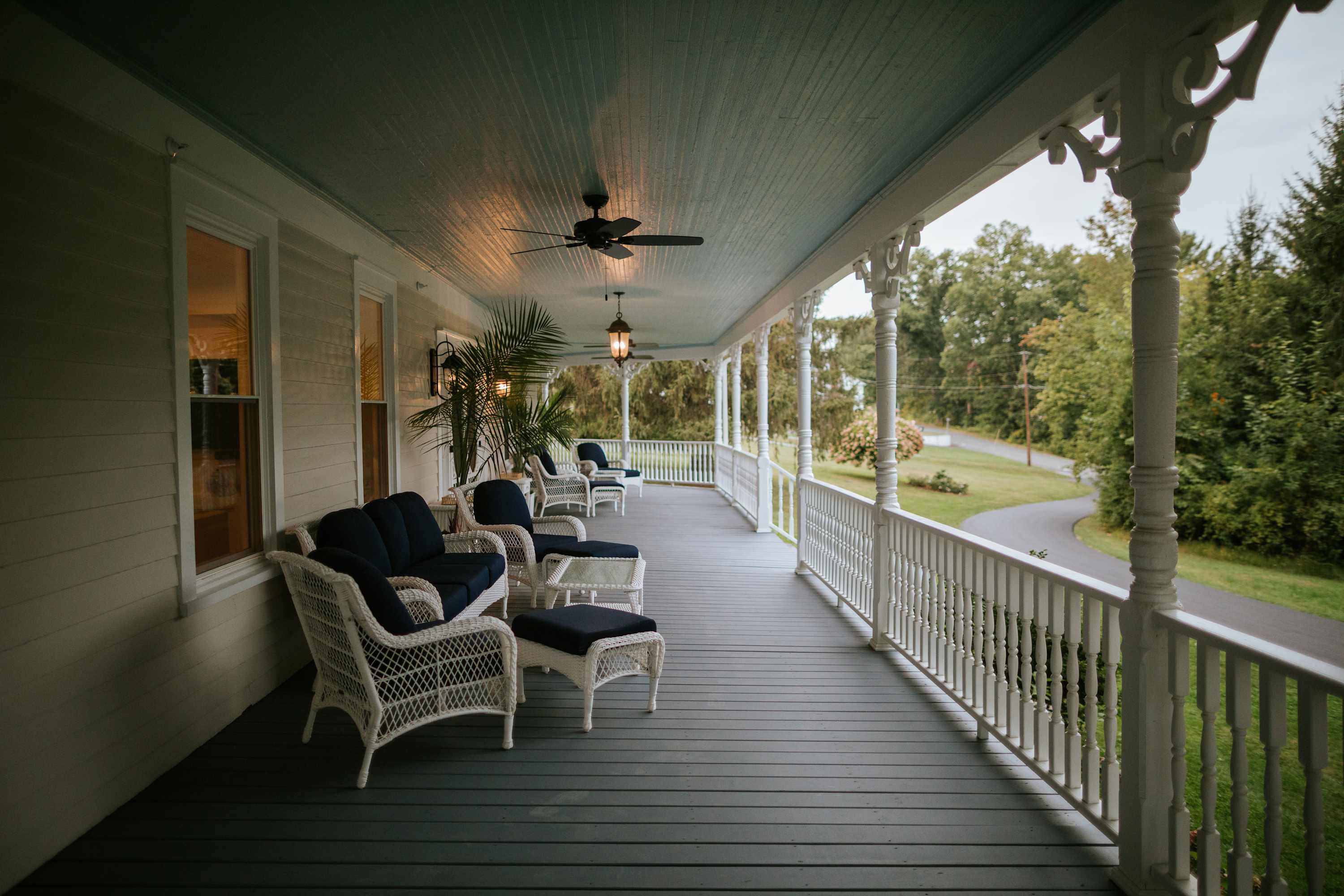 A long, inviting wrap-around porch with a light blue ceiling, white wicker seating with dark blue cushions, and decorative white railings. Ceiling fans and hanging lanterns provide comfort and ambiance.
