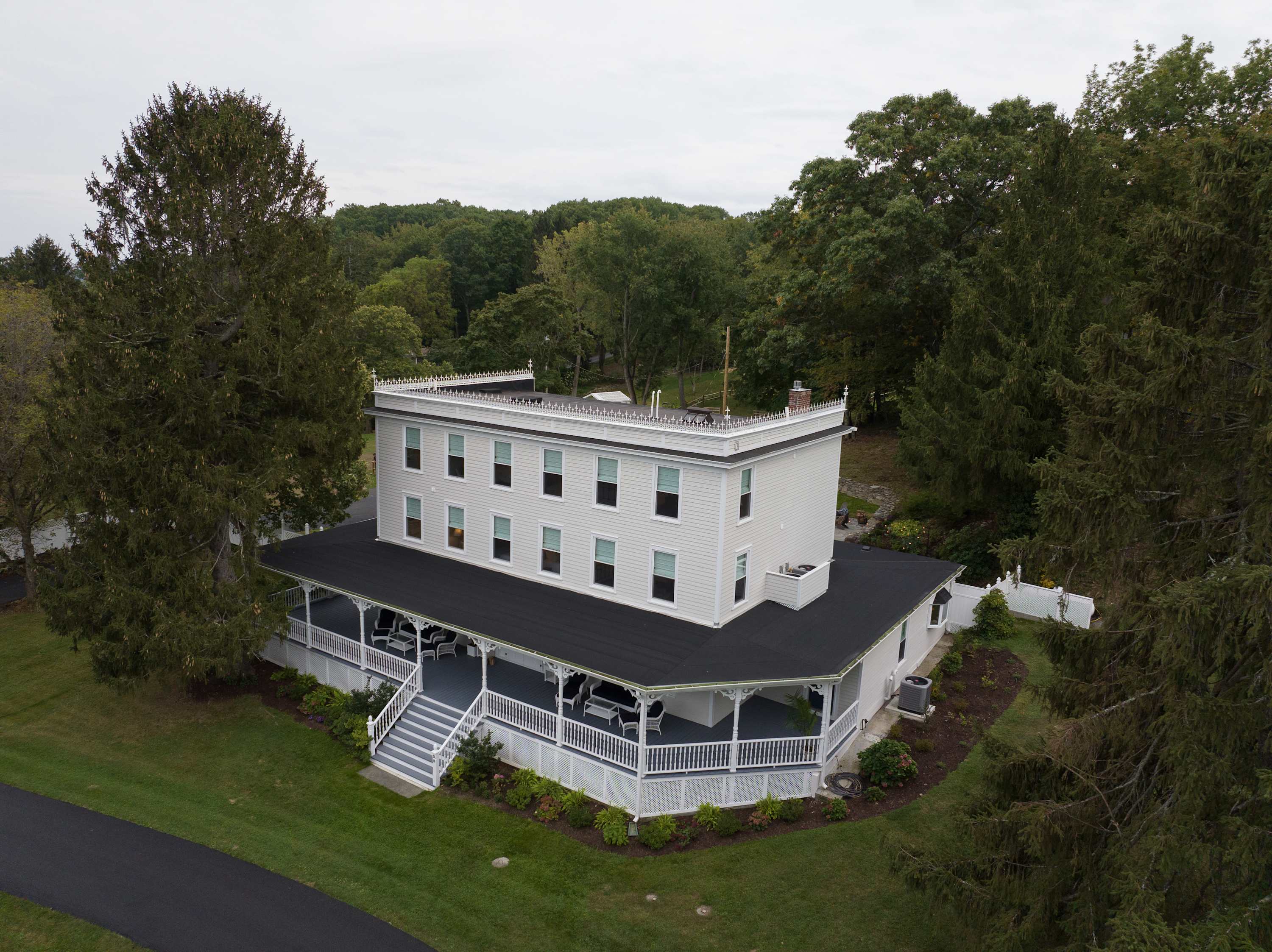 An aerial view of a large, white multi-story house with a dark roof and an expansive wrap-around porch. The property is surrounded by lush green trees and a well-maintained lawn, with a paved driveway leading up to the house.