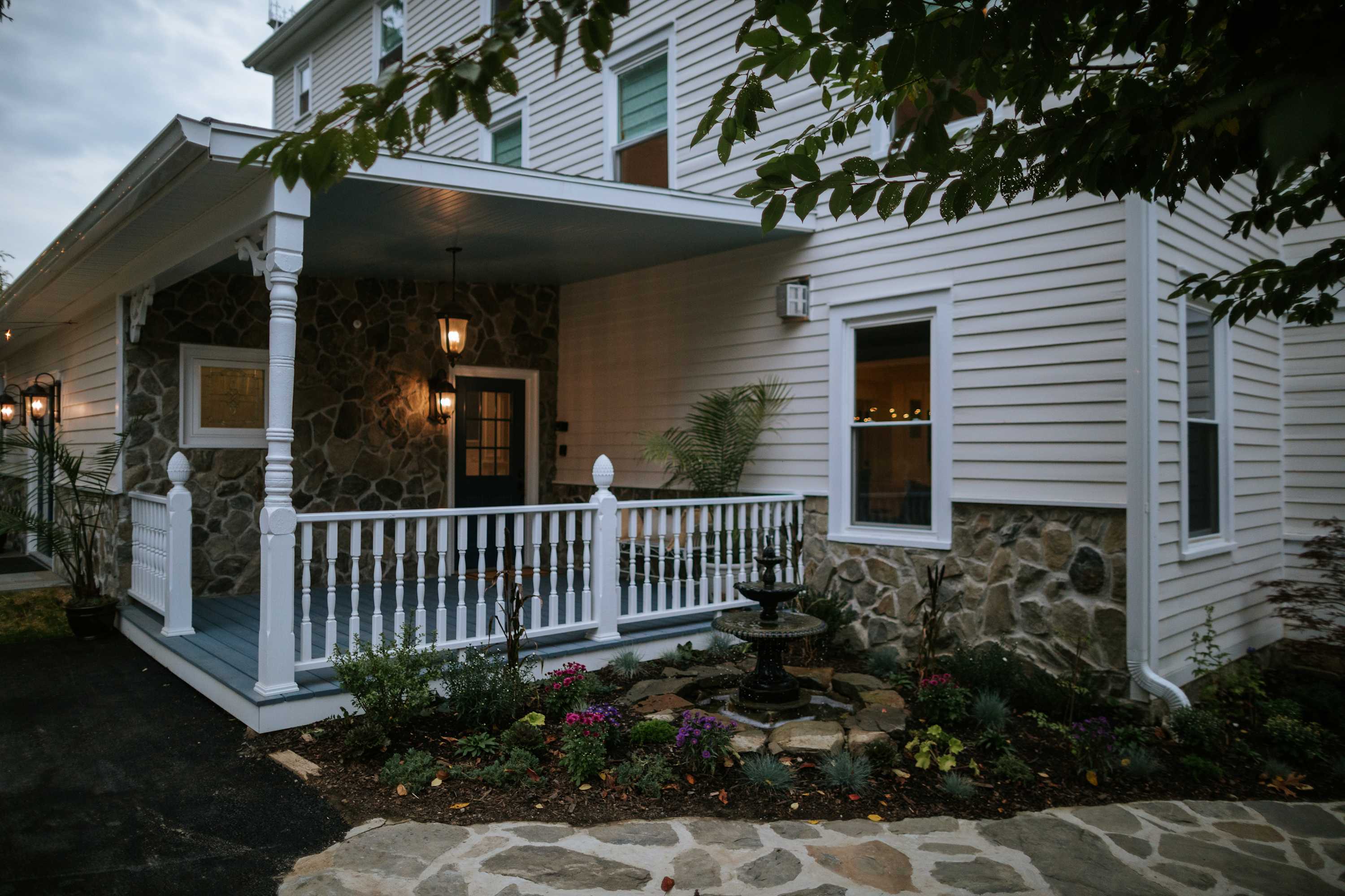 A welcoming front entrance with a covered porch supported by white columns and railings. The lower part of the house features stone cladding, while the upper part has white siding. A small fountain and garden beds enhance the curb appeal.