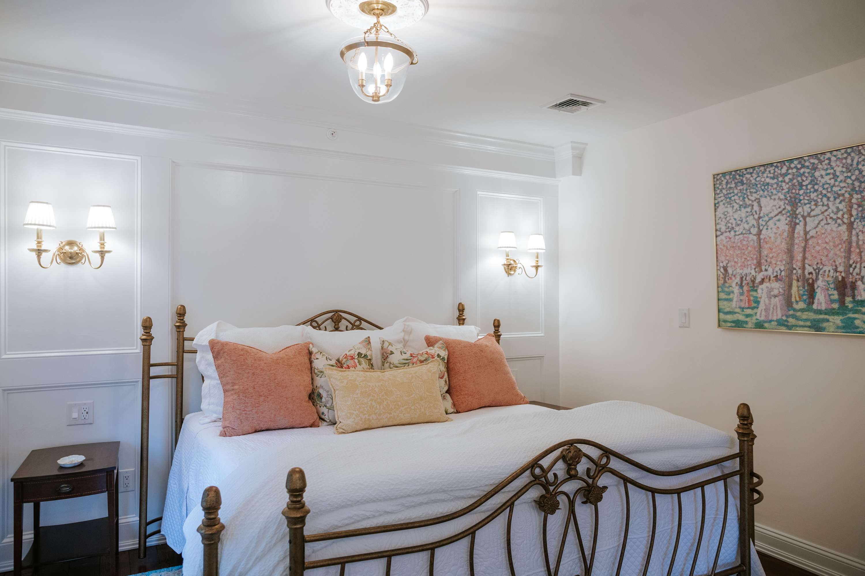 A cozy bedroom featuring a brass-colored metal bed frame with a white comforter and decorative pillows in coral and yellow. The walls are white with decorative molding, and two wall sconces provide ambient lighting.