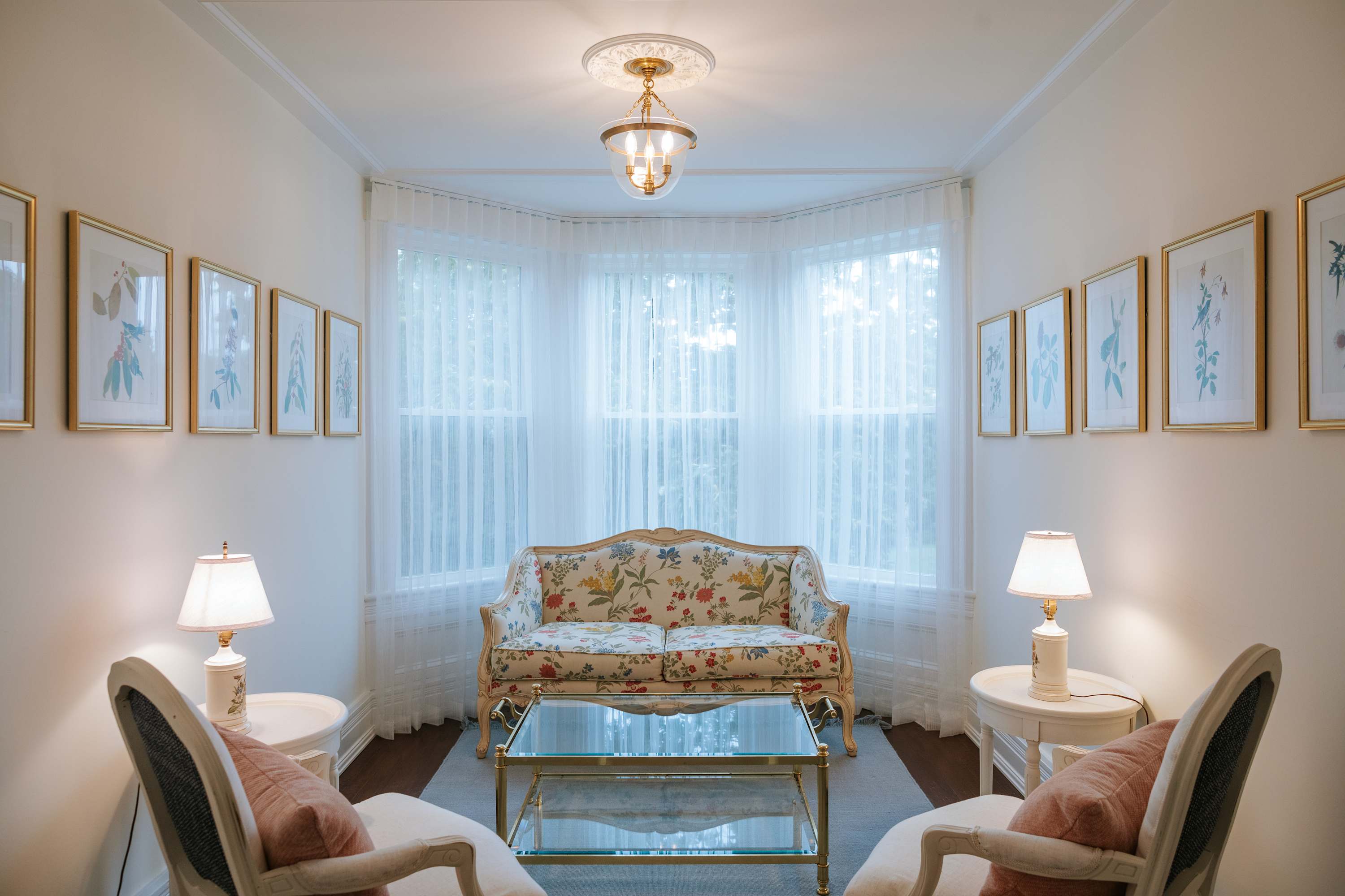 An elegant living space with a floral patterned sofa and two white armchairs, arranged around a glass coffee table on a light rug. A bay window with sheer white curtains and multiple framed botanical prints adorn the walls.