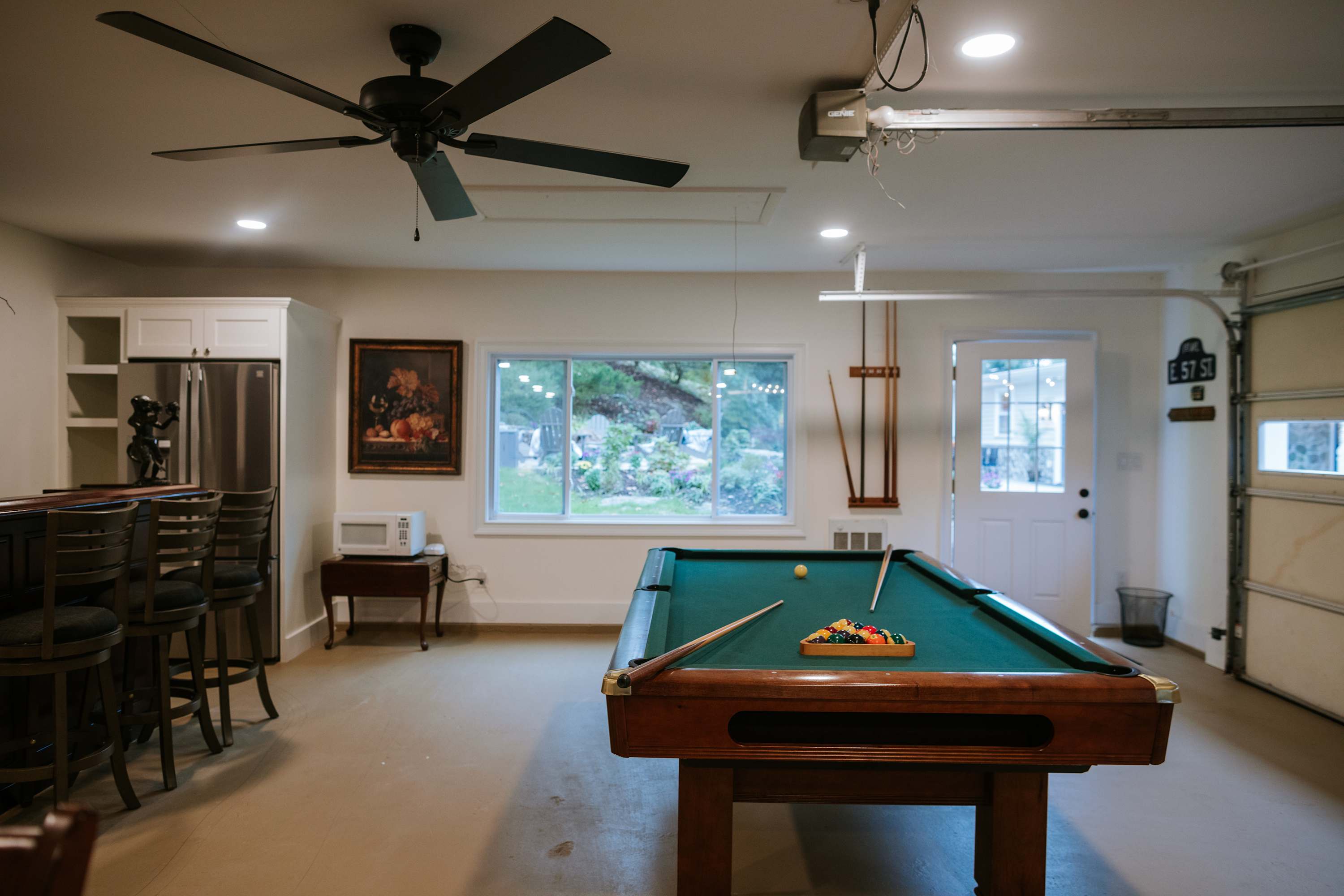 A spacious game room features a green felt pool table with cues and balls, a dark wood bar with stools, and a large window overlooking a lush garden. A ceiling fan and recessed lighting illuminate the light-colored walls and floor.