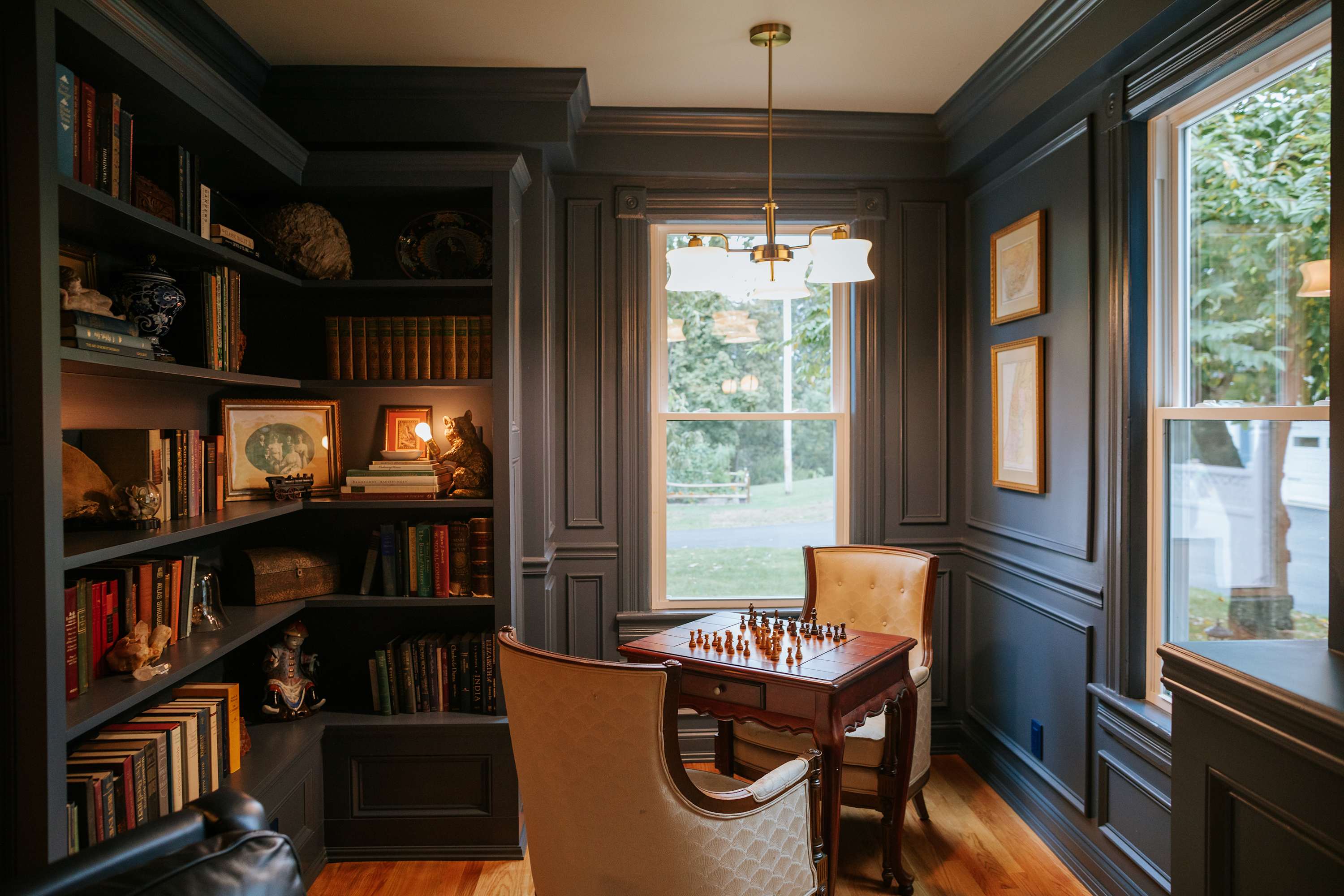 Cozy study room with dark paneled walls, built-in bookshelves filled with books, and a small wooden chess table with two upholstered chairs. A window overlooks a green outdoor area.