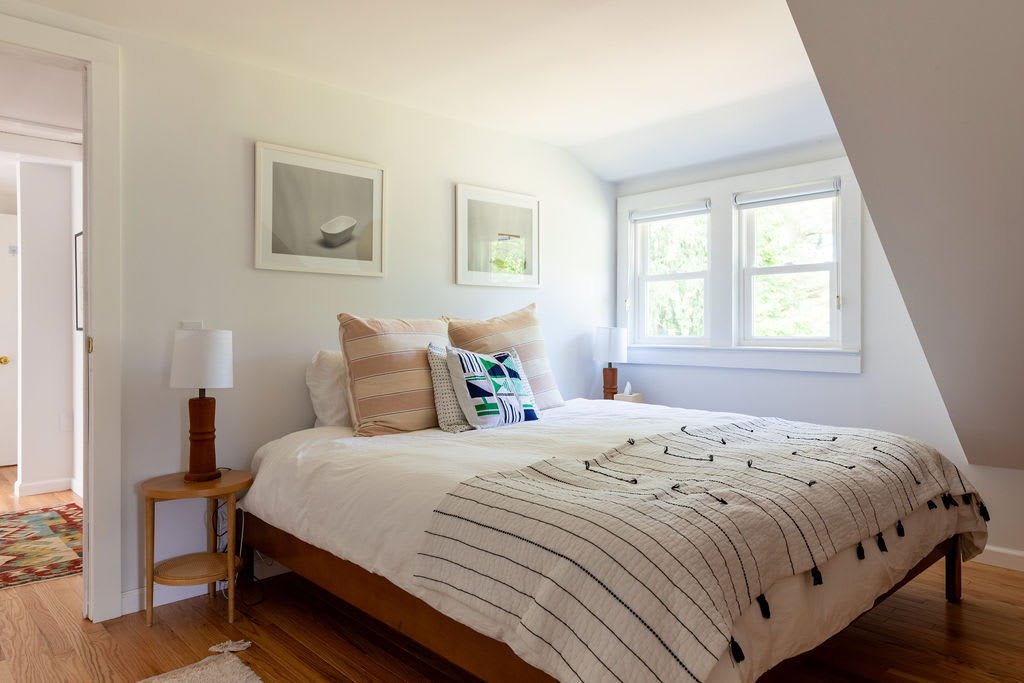 A cozy bedroom with a queen-sized bed dressed in white linens and a striped throw, flanked by wooden nightstands with lamps, under two framed artworks and a window.