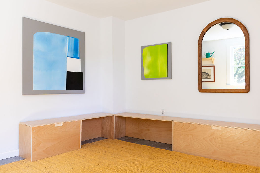 A minimalist room with white walls and a yellow textured rug, featuring custom light wood built-in benches and storage along two walls, adorned with modern abstract art and an arched wooden mirror.