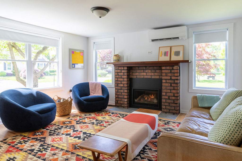 A cozy living room with light walls, hardwood floors, and a brick fireplace with a wooden mantel, furnished with a brown leather sofa, two blue barrel chairs, and a colorful geometric area rug.