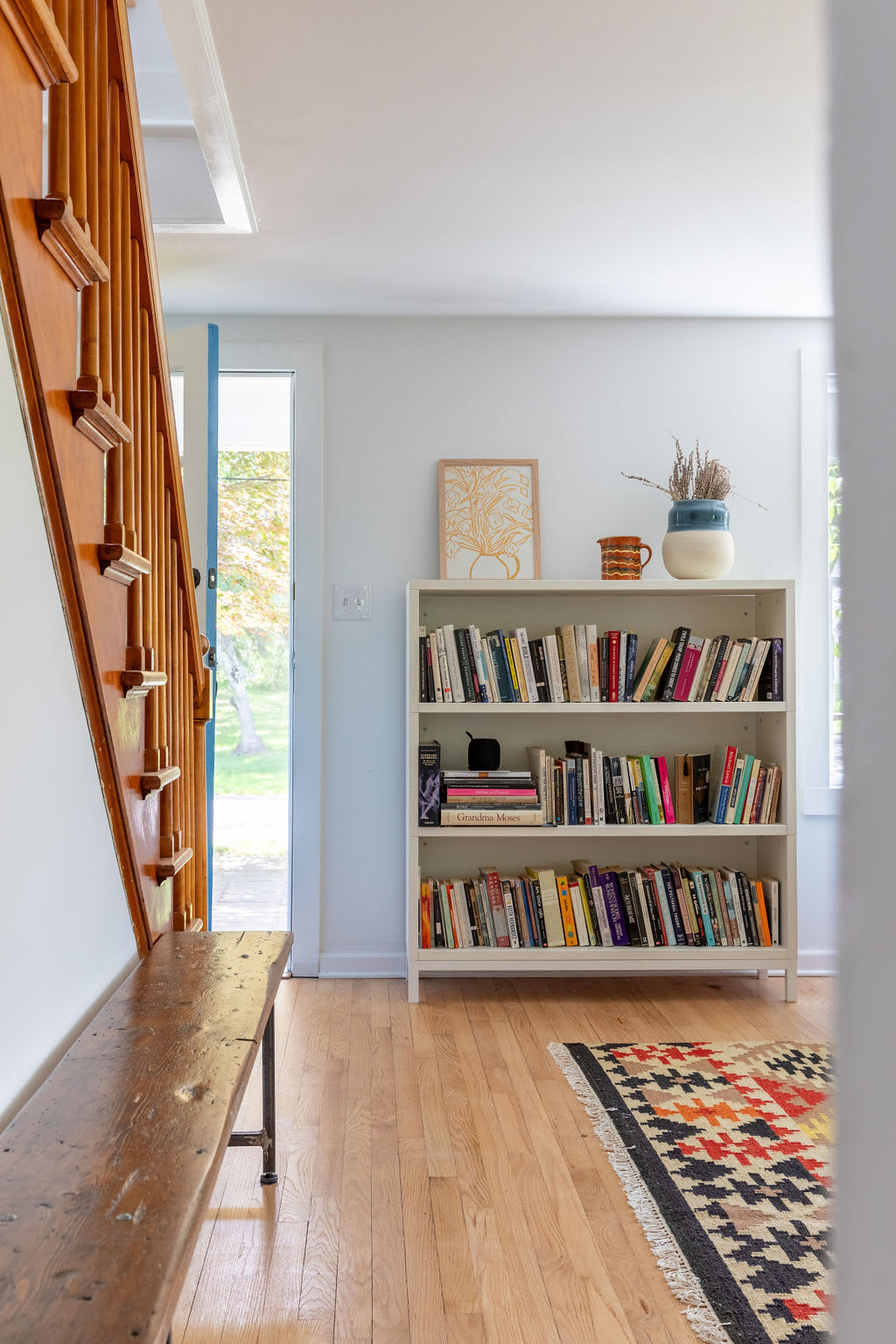 A bright foyer features a wooden staircase with a banister, a long rustic wooden bench, and a white bookshelf filled with books, all on light hardwood floors.