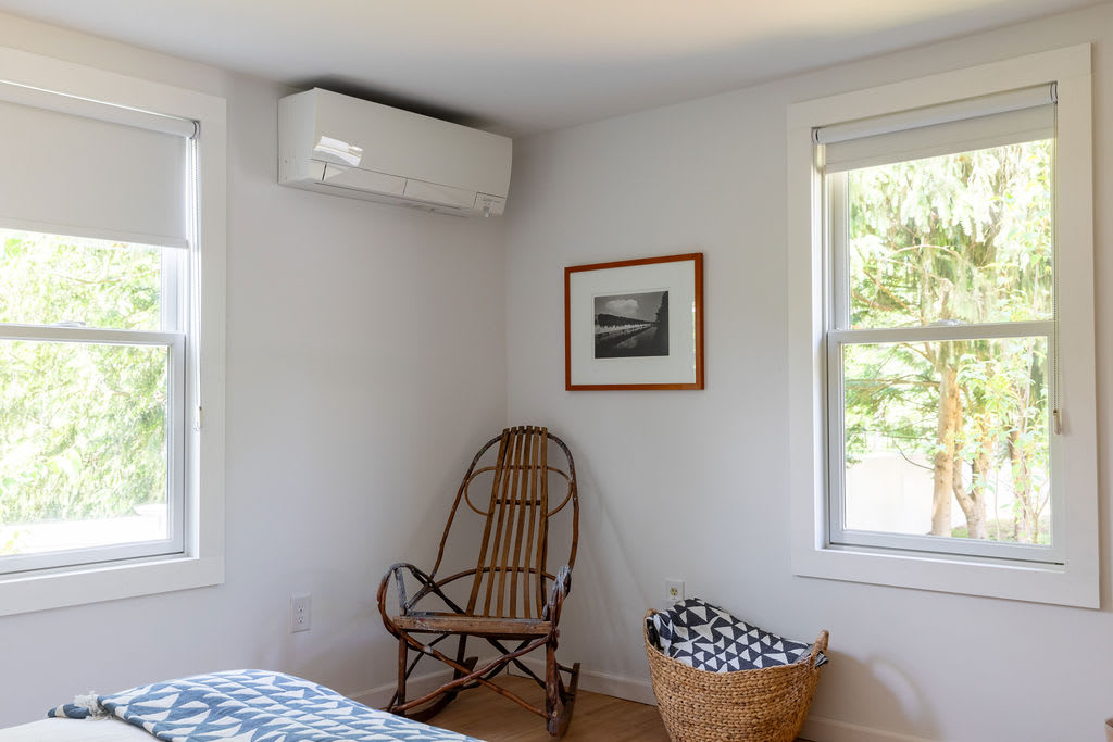 A bright bedroom with white walls and hardwood floors, featuring a modern wall-mounted air conditioning unit, a unique wooden rocking chair, and two large windows offering views of lush greenery.