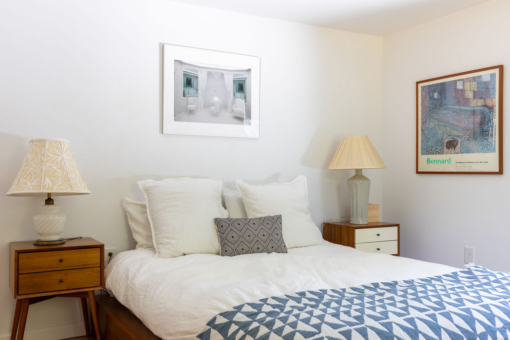 A bright bedroom featuring a queen bed with white bedding and a blue geometric blanket, flanked by two wooden nightstands with lamps. A large framed artwork hangs above the bed, and a colorful poster is on the adjacent wall.