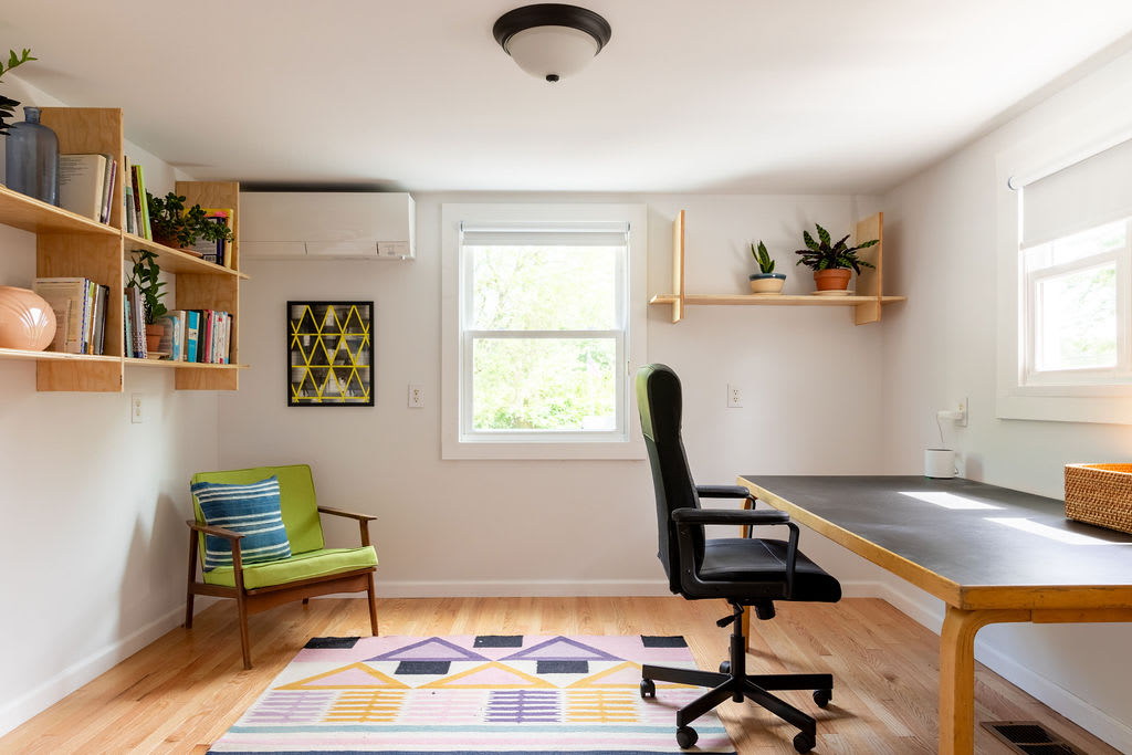 Bright home office with light hardwood floors and white walls, featuring a large desk with a black office chair and a green accent chair. Floating wooden shelves display books and plants, with a mini-split AC unit mounted on the wall.