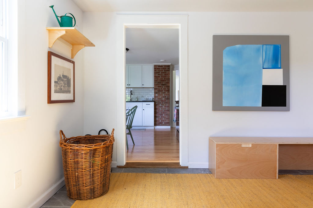 A bright entryway or hallway with white walls and a yellow textured rug, featuring a large wicker basket, a wooden shelf with a watering can, and an open doorway leading into the kitchen area.
