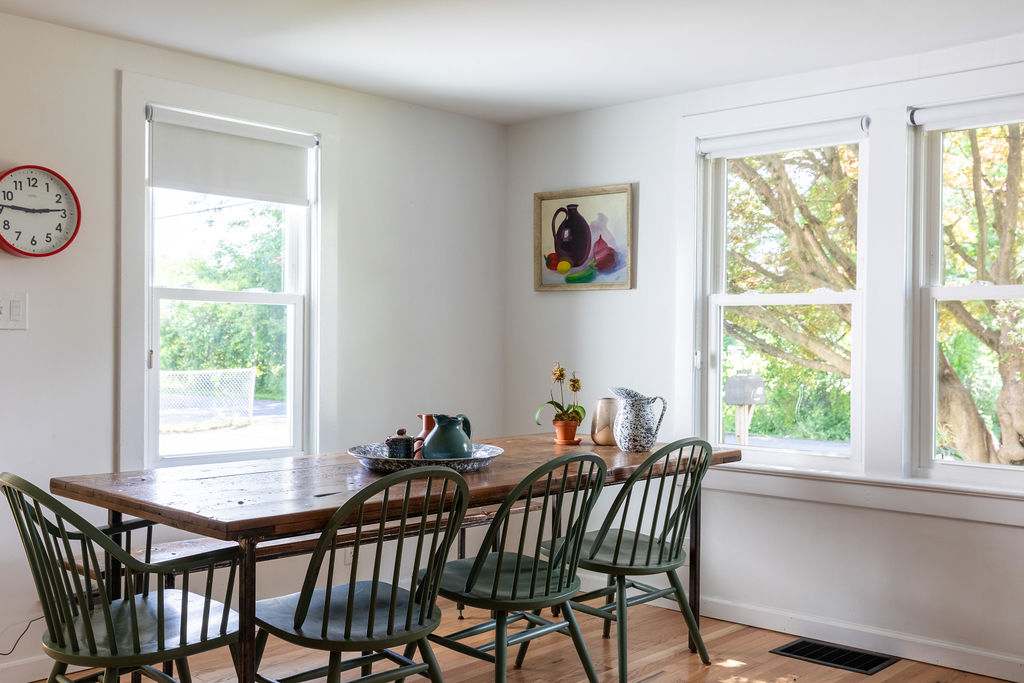 A sunlit dining area with a rustic wooden table and four dark green Windsor-style chairs, positioned by large windows, with white walls and hardwood floors, and a vintage wall clock.