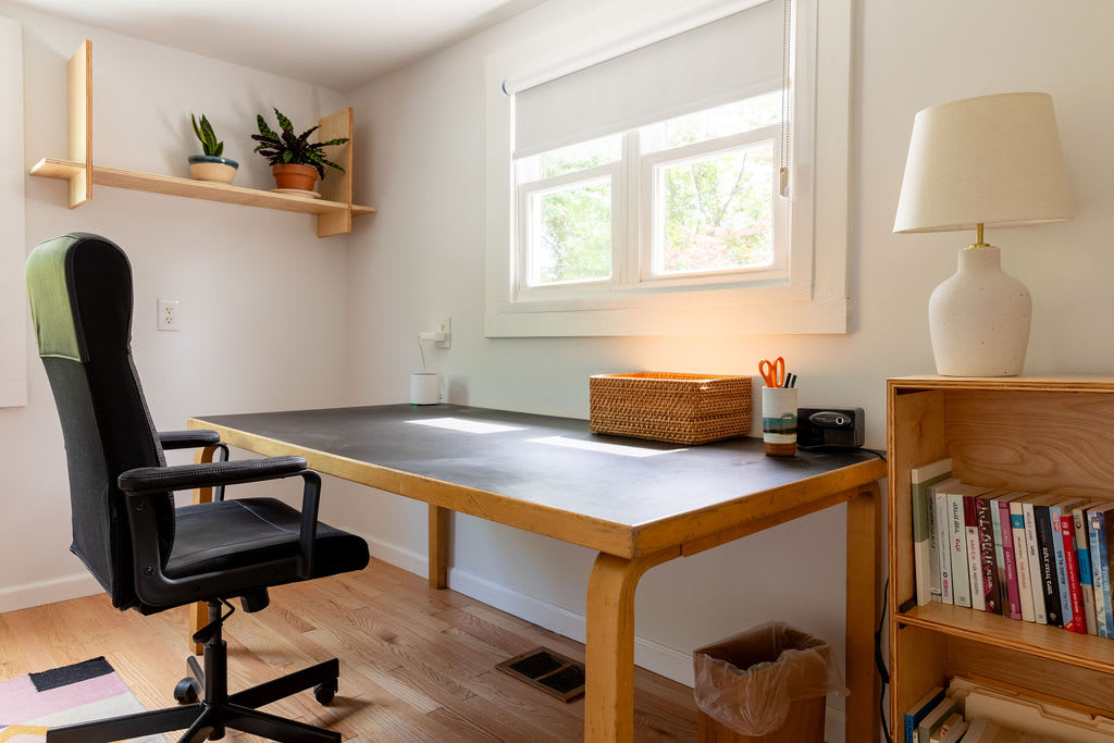 A dedicated office space features a large wooden desk with a black top, a black office chair, and a window providing natural light, with a small wooden bookshelf to the side.