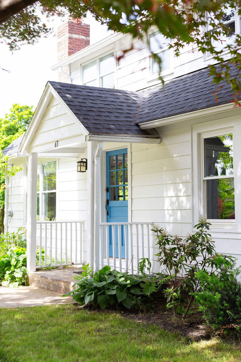 A side view of the white house highlights its blue front door and a covered porch with white railings. The home features a dark shingled roof and a prominent brick chimney, set against a backdrop of green lawn and garden beds.