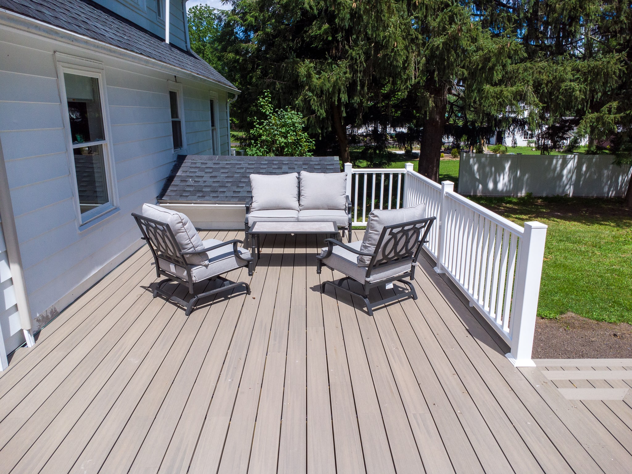 A spacious outdoor deck with light-colored composite decking, featuring a white railing and a modern gray outdoor seating set including a sofa and two armchairs with light cushions, surrounded by lush green trees.