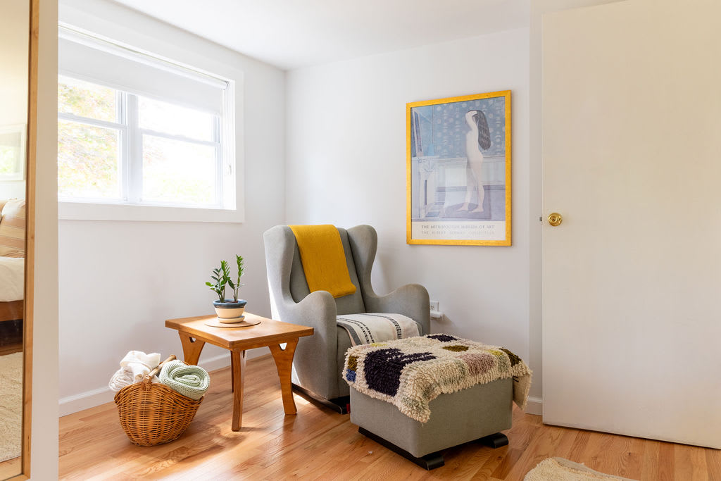 A bright reading nook features a grey upholstered armchair with a yellow throw, a matching ottoman, a small wooden side table, and a large framed poster on the wall.