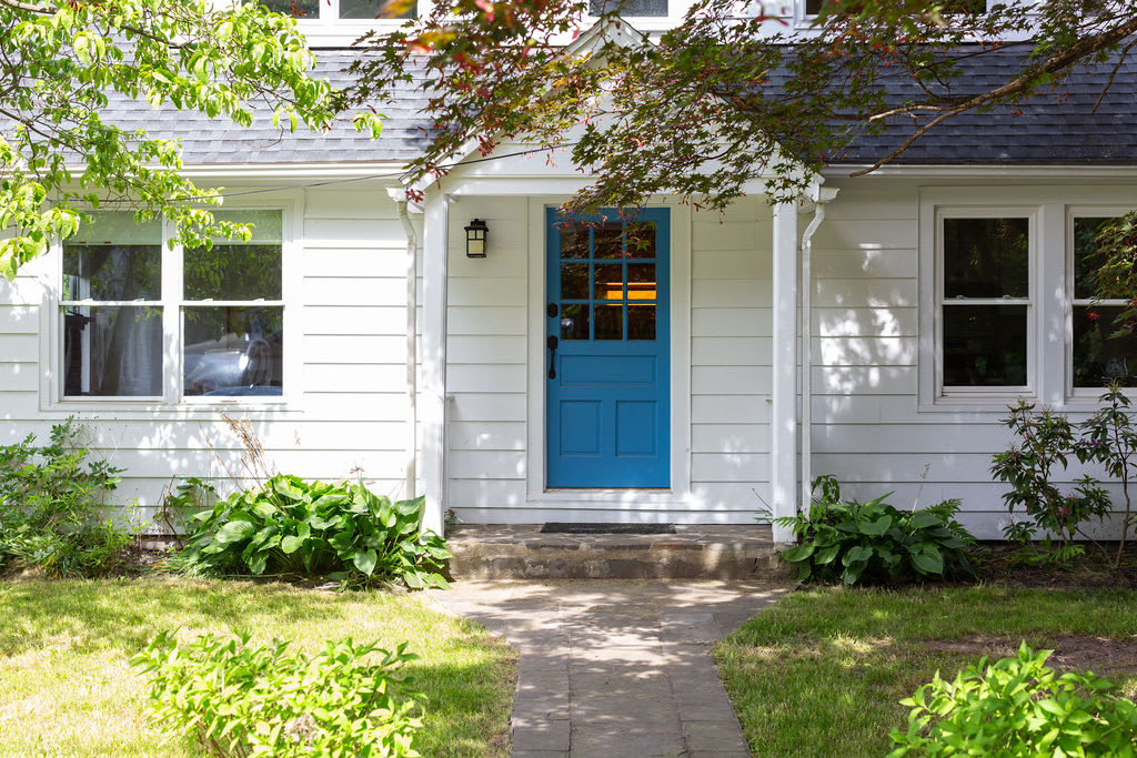The front facade of a charming white house features a vibrant blue front door with glass panes, flanked by large windows. A small covered porch with steps leads to the entrance, surrounded by lush green grass and mature landscaping.