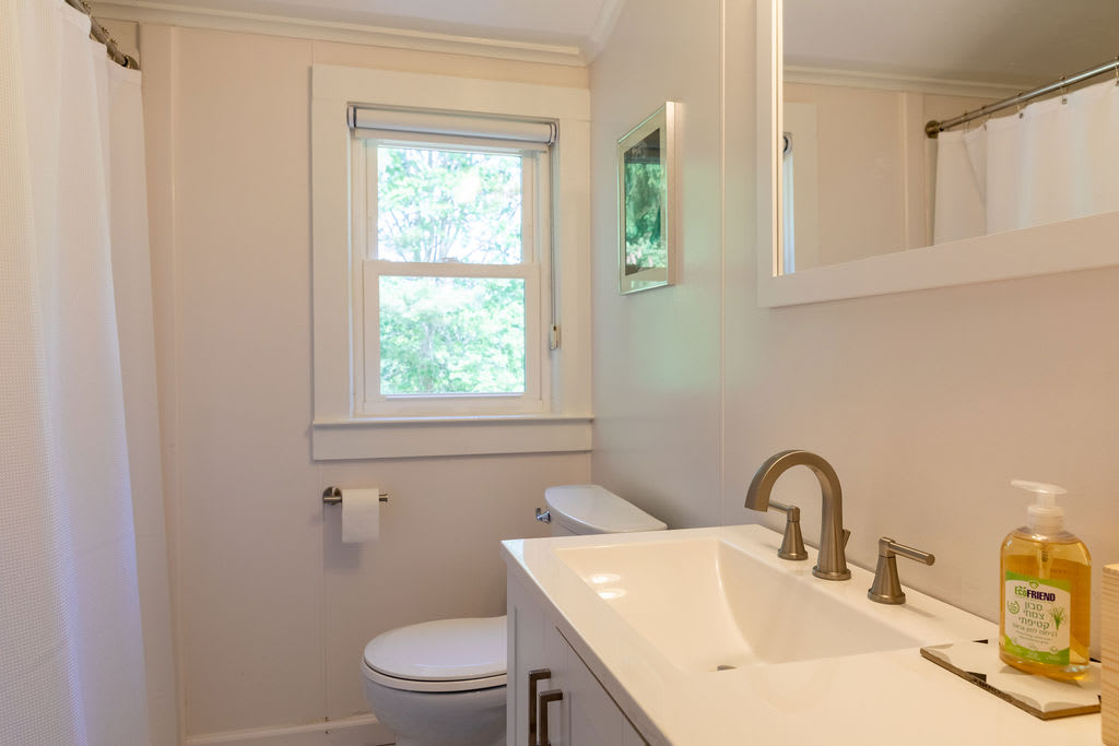 A clean bathroom features a white vanity with a modern sink and faucet, a toilet, and a window, with a shower curtain visible on the left.