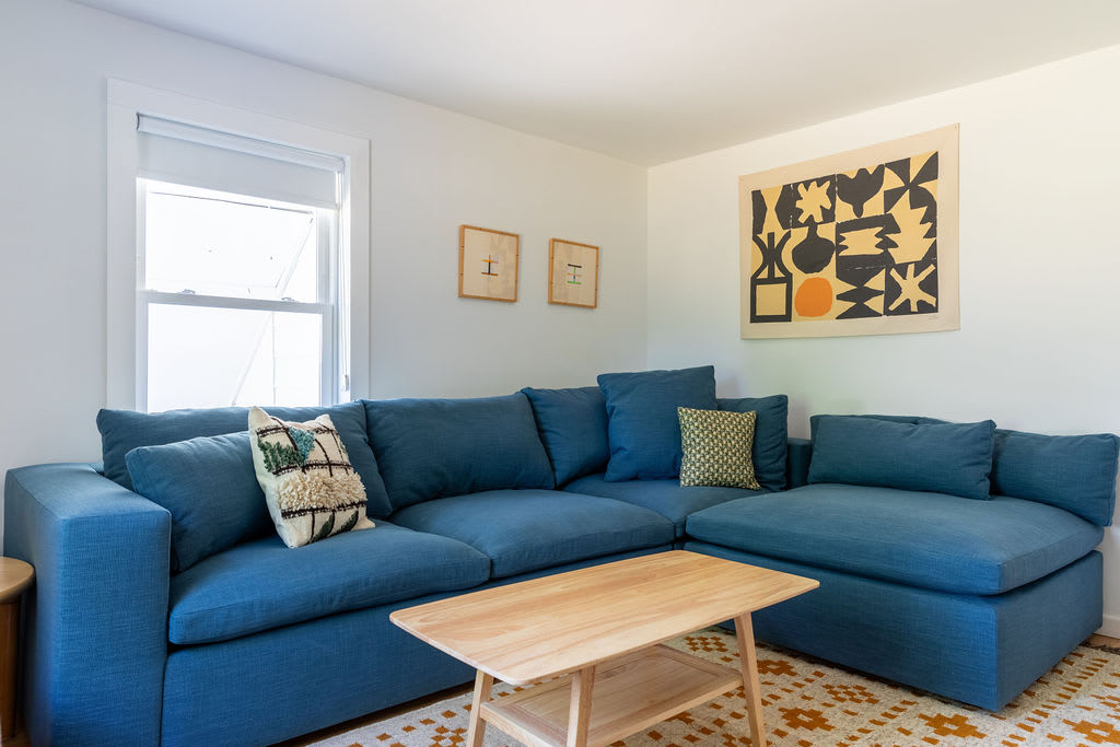 A cozy living area featuring a large blue sectional sofa with various throw pillows, centered around a light wood coffee table on a patterned rug. White walls and a window provide natural light, with abstract art hanging above the sofa.