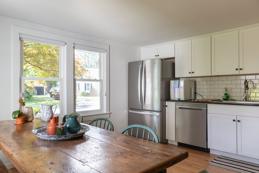 A bright kitchen and dining area with white cabinets and stainless steel appliances, featuring a large rustic wooden dining table with light blue chairs, positioned next to two large windows overlooking the outdoors.