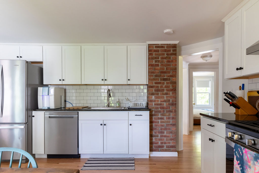 A modern kitchen featuring white shaker cabinets, black countertops, and a white subway tile backsplash, with stainless steel appliances including a refrigerator and dishwasher, and an exposed brick column adding a rustic touch.