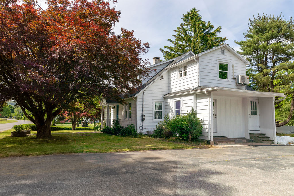 The white-sided house features multiple windows and a covered entrance, surrounded by lush green grass and mature trees, including a prominent red-leafed tree.