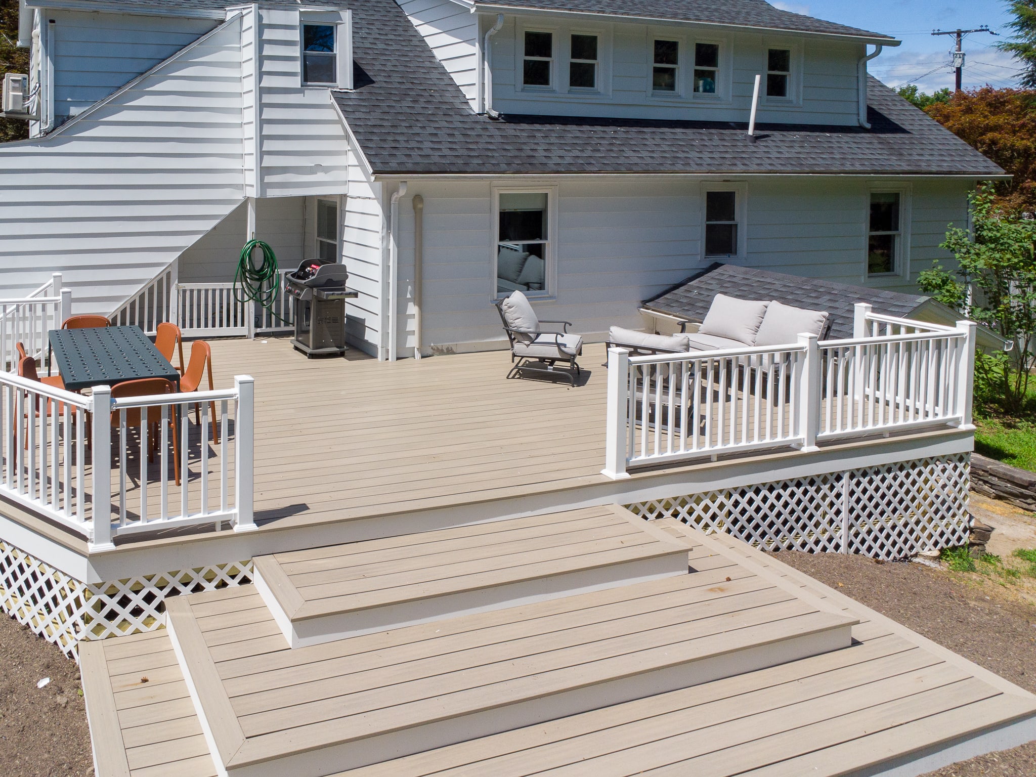 A spacious multi-level composite deck with white railings extends from the back of the house, furnished with an outdoor dining set, a lounge sofa, and a BBQ grill, overlooking a grassy backyard.
