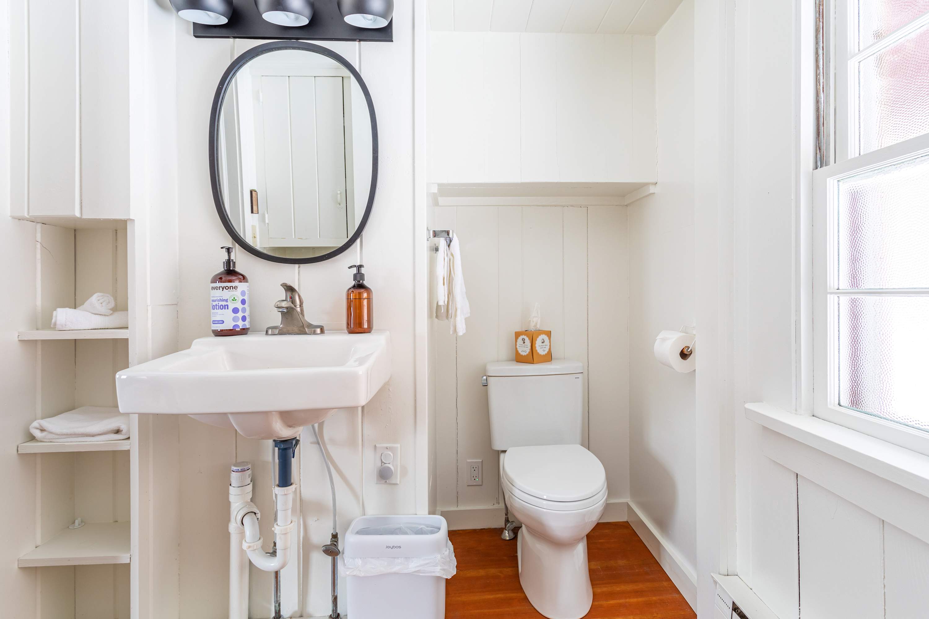 Bright bathroom with white walls and beadboard, featuring a pedestal sink with a round mirror, a toilet, and open shelving with towels and toiletries.