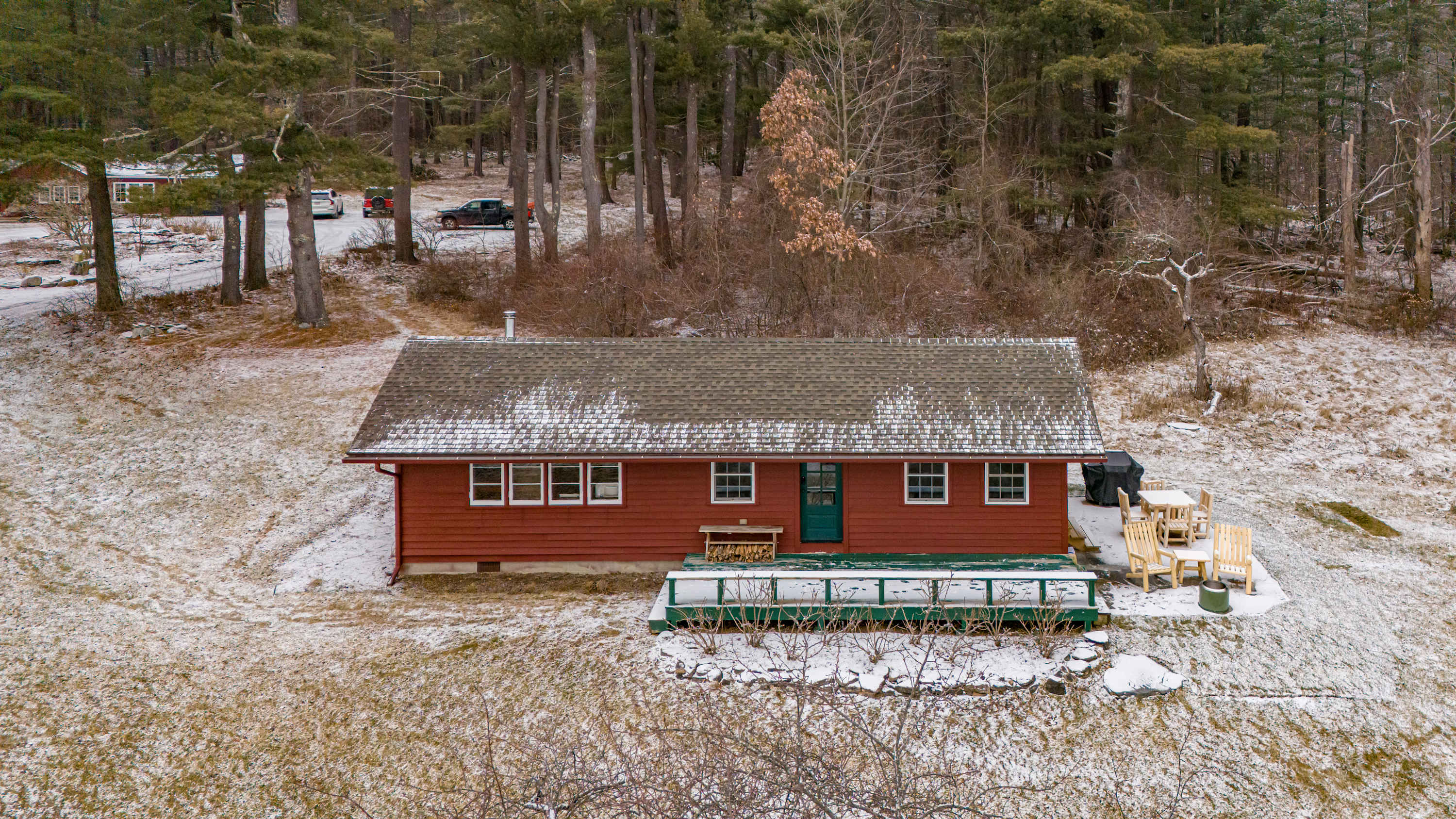 Elevated aerial view of the red cabin's front, showcasing its green-railed deck and an adjacent patio with wooden outdoor furniture. The property is set against a backdrop of snow-dusted ground and a dense pine forest.