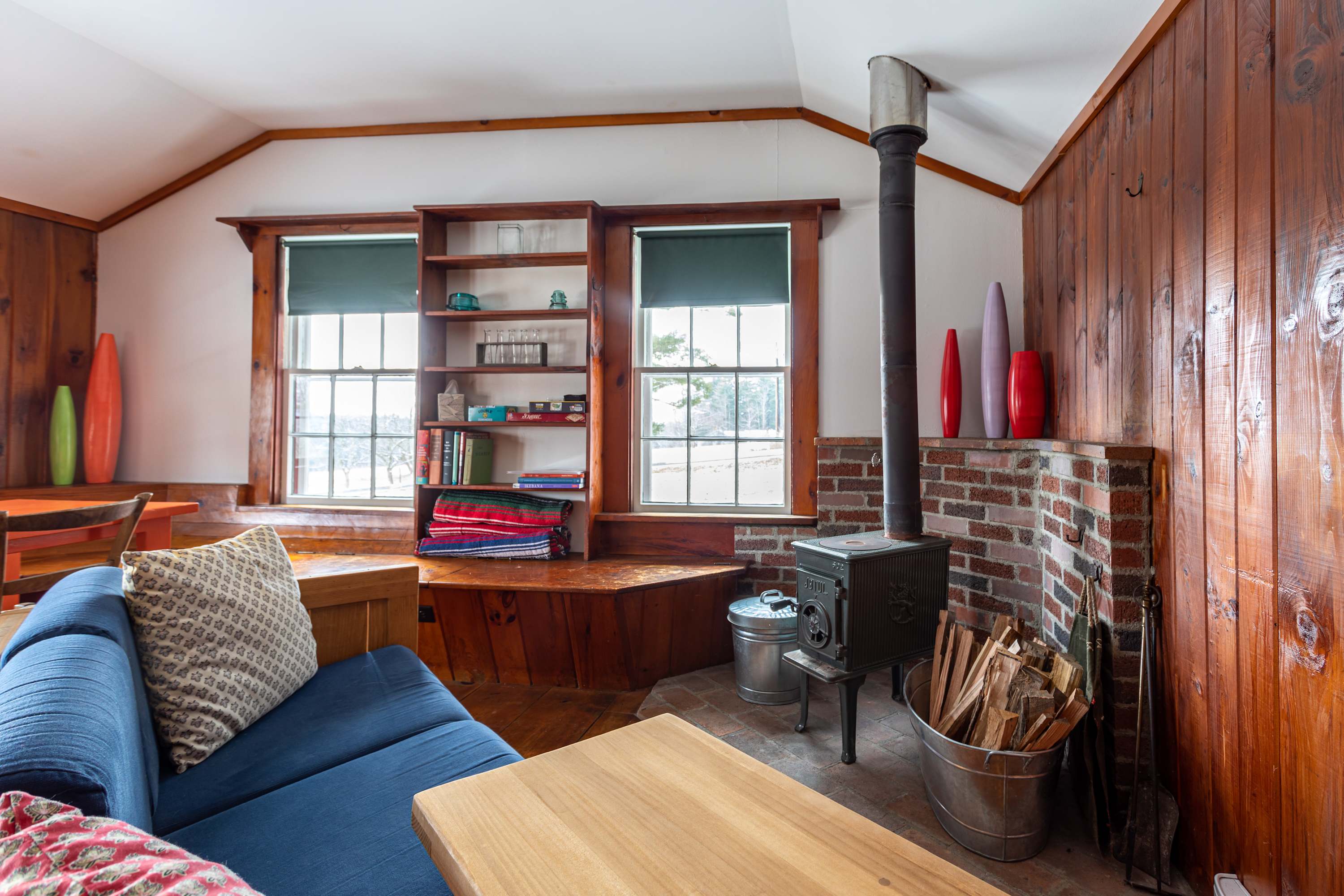 A warm living room with knotty pine walls and a vaulted ceiling, centered around a black wood-burning stove on a brick hearth, with a blue sofa and built-in shelving visible.