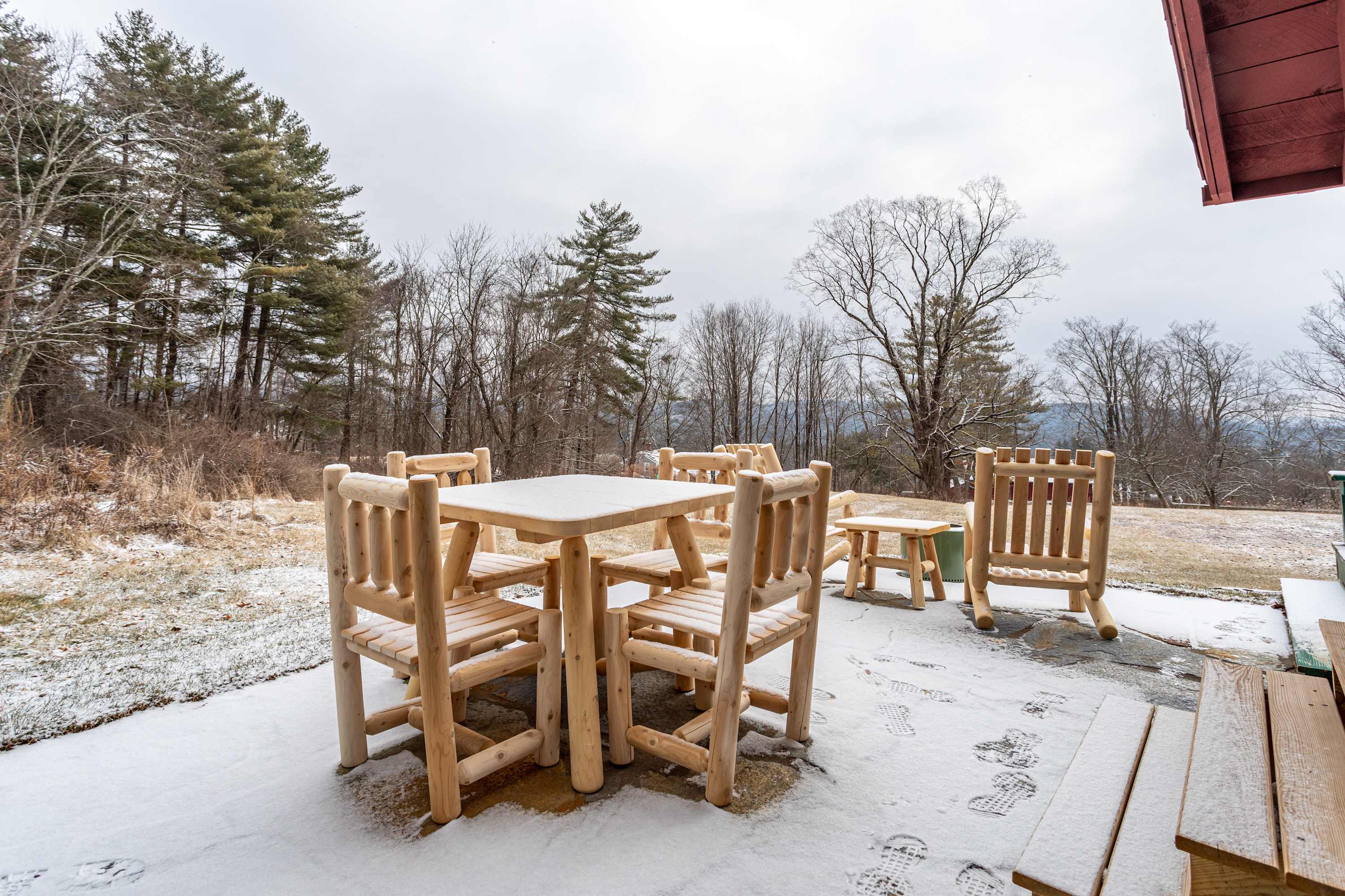 Snowy outdoor patio with a rustic wooden dining table and chairs, surrounded by a winter landscape with bare trees and a distant red building.