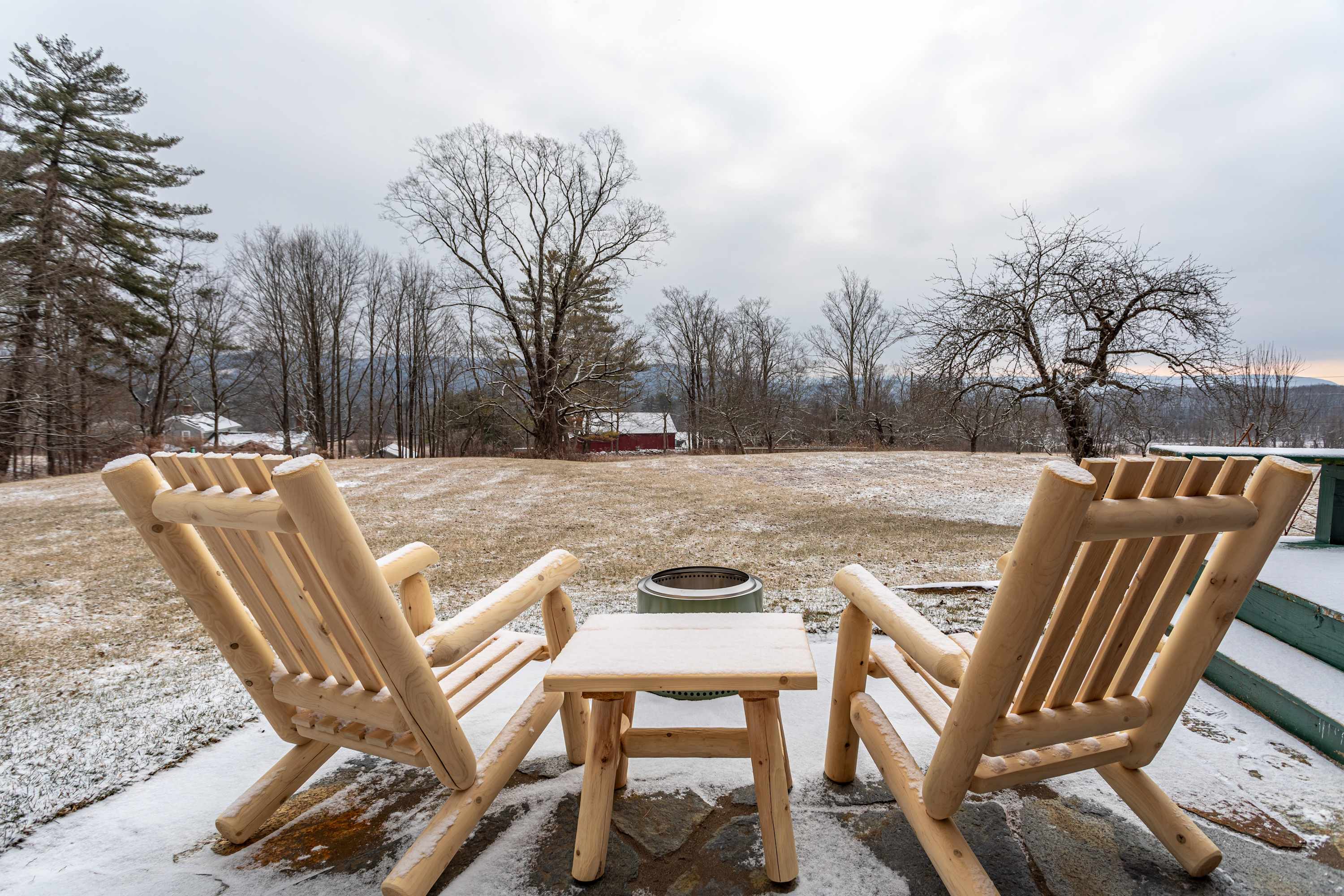 Outdoor patio covered in light snow, featuring two rustic wooden chairs facing a metal fire pit, with a scenic view of a winter field and distant trees.