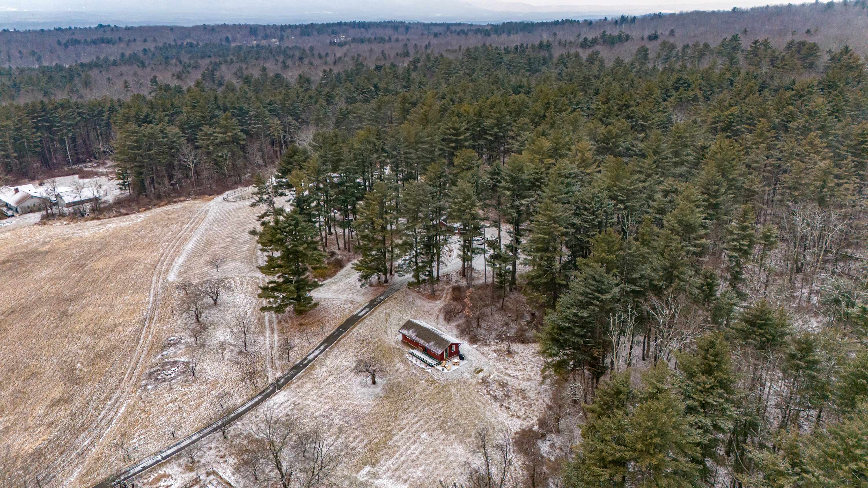 High aerial view showcasing a red cabin nestled within a vast, snow-dusted forest, with a winding dirt road leading to it. An expansive open field stretches to the left, under a cloudy sky.