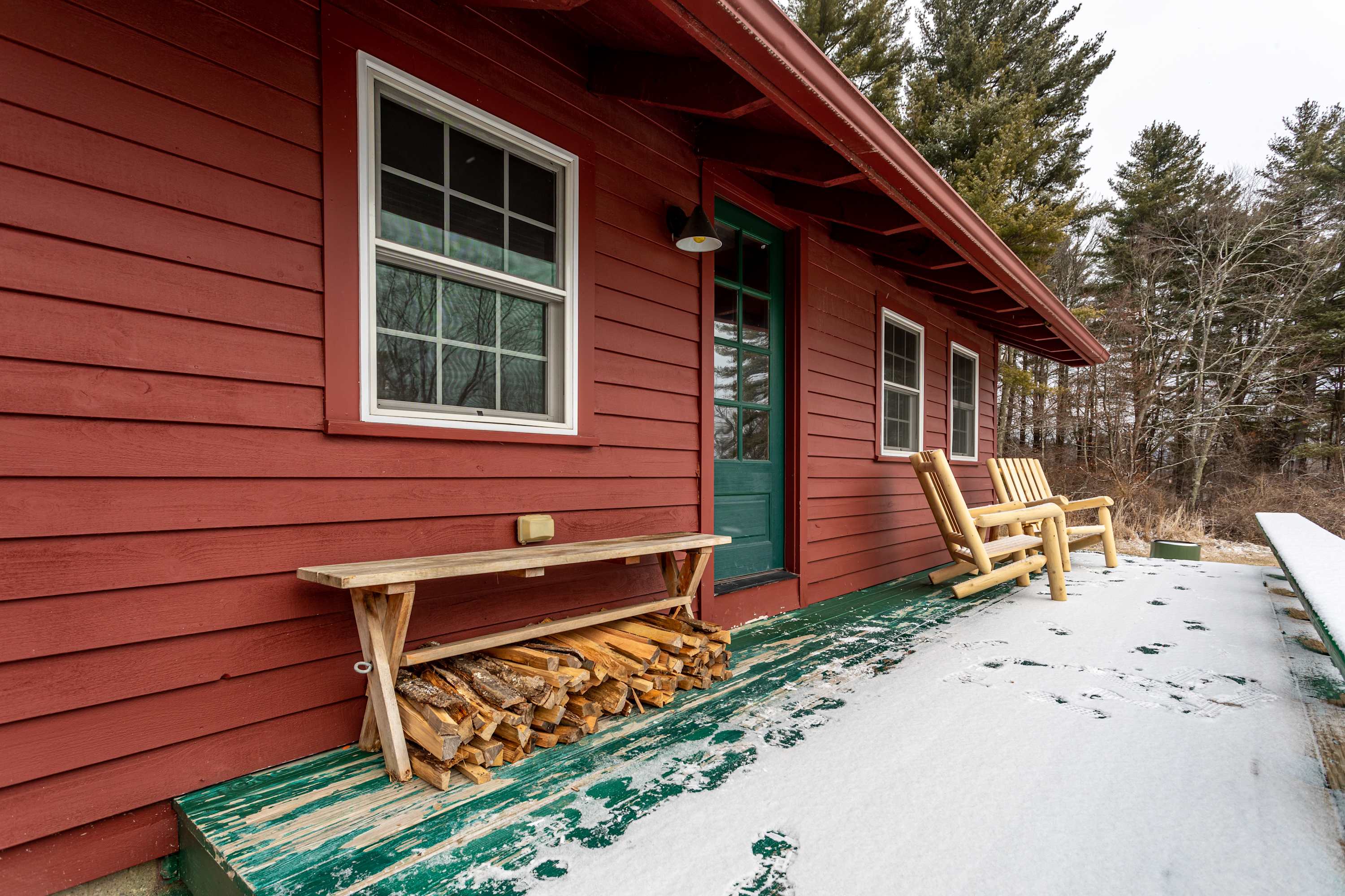 Exterior view of a red cabin's snowy porch, featuring a stack of firewood under a bench, a green entry door, and rustic wooden chairs, surrounded by a winter scene.