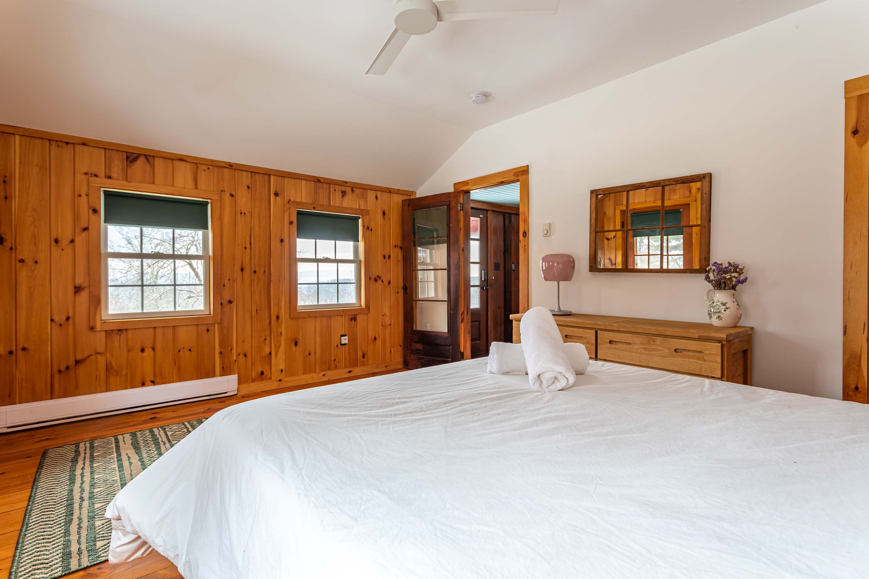 Cozy bedroom featuring a queen bed with white linens, a wooden dresser with a mirror, and multiple windows offering views of the snowy outdoors, all set against warm wood paneling.
