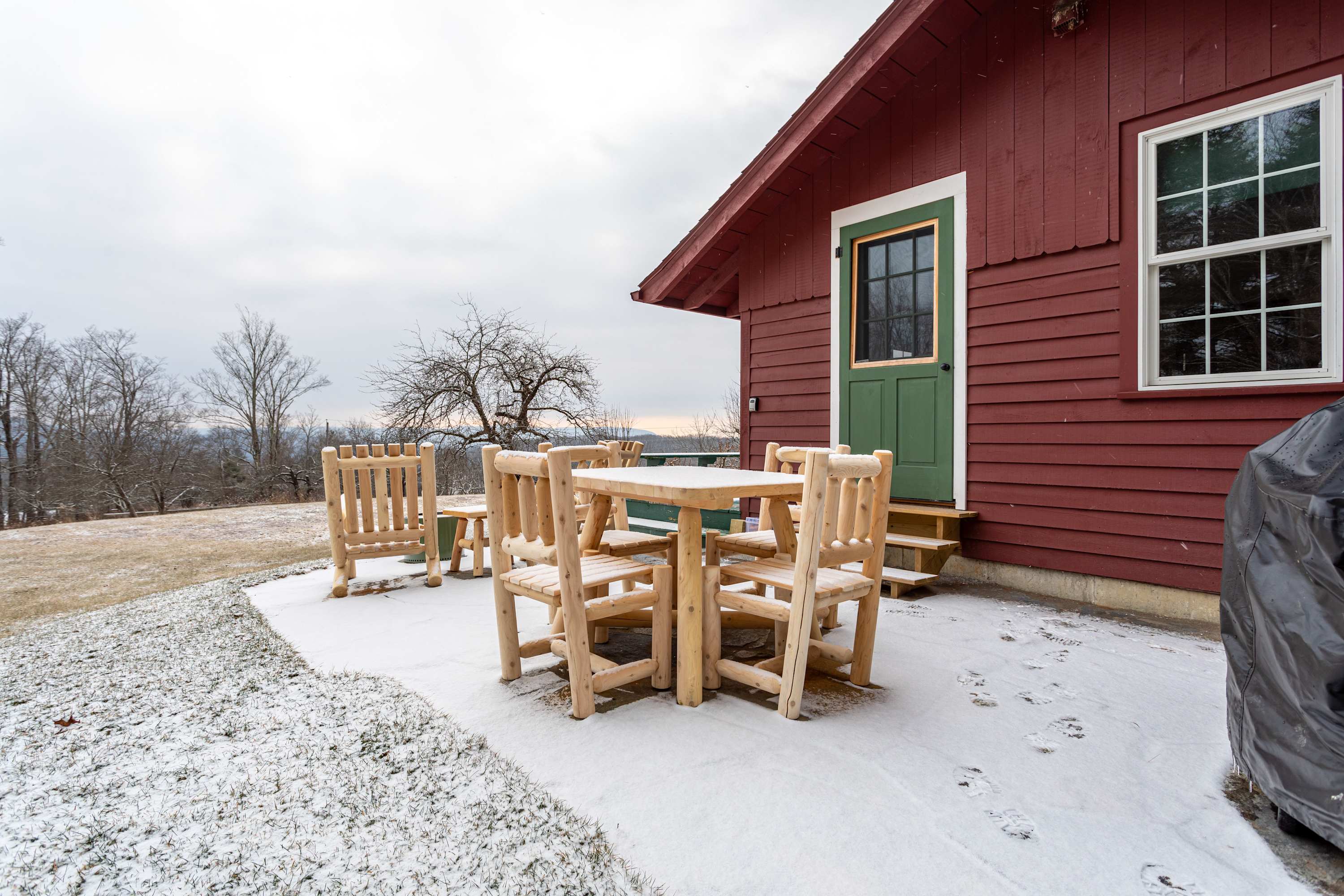The red cabin's concrete patio and green deck in winter, featuring a light wood outdoor dining set and a grill cover, all lightly dusted with snow and surrounded by a sparse, wintry landscape.