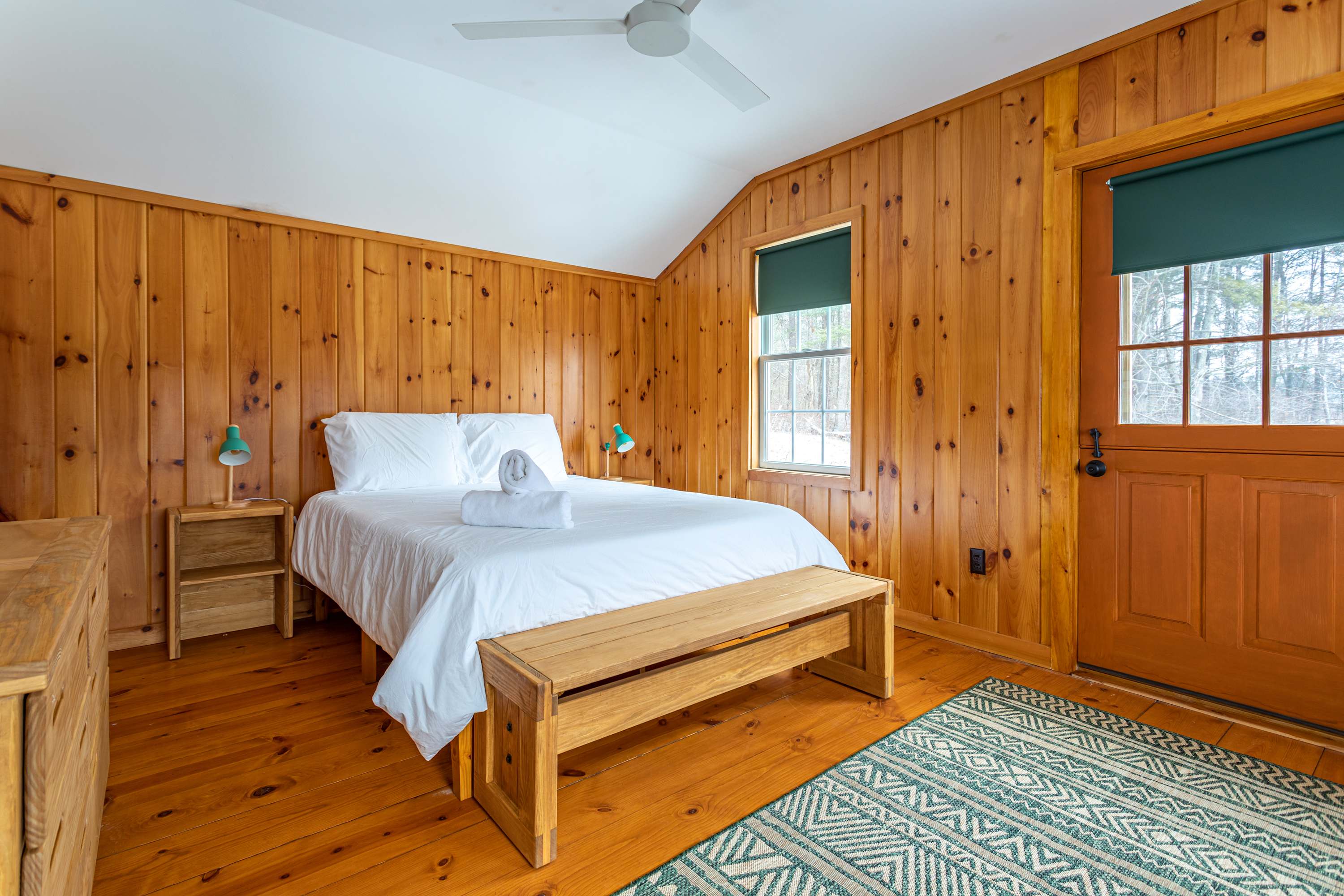 A warm bedroom with light wood paneling on all walls and polished hardwood floors, featuring a queen-sized bed with white bedding, two wooden nightstands, and a window with a dark green blind. A wooden bench sits at the foot of the bed, and a patterned green rug is on the floor.