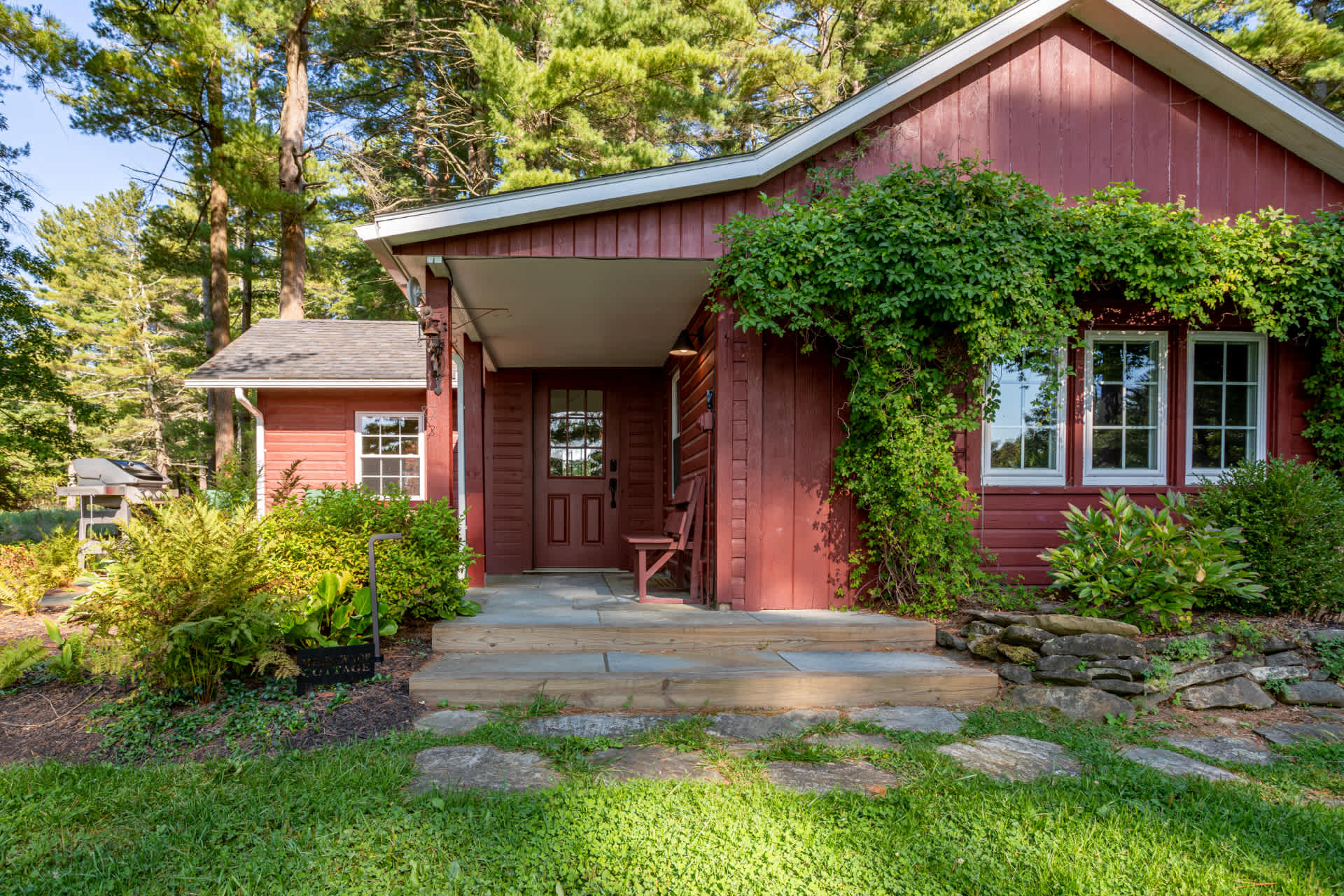 Charming red cottage with a welcoming front entrance, featuring a small porch with stone steps and lush green ivy climbing the walls. The house is nestled among mature trees and surrounded by well-maintained landscaping.
