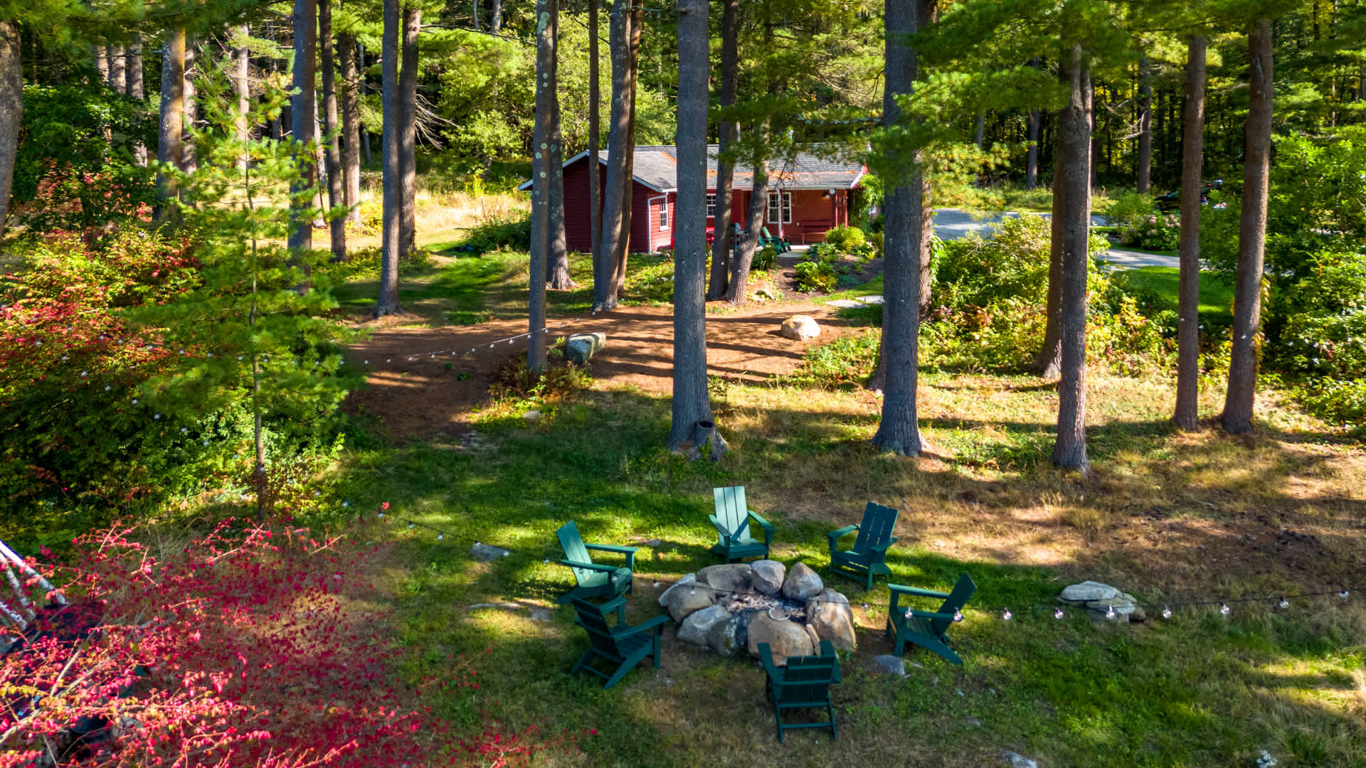 An aerial view shows a circular stone fire pit surrounded by six green Adirondack chairs on a grassy lawn, with a red cottage nestled among tall pine trees in the background. Red foliage and green bushes frame the scene, suggesting an autumn setting.